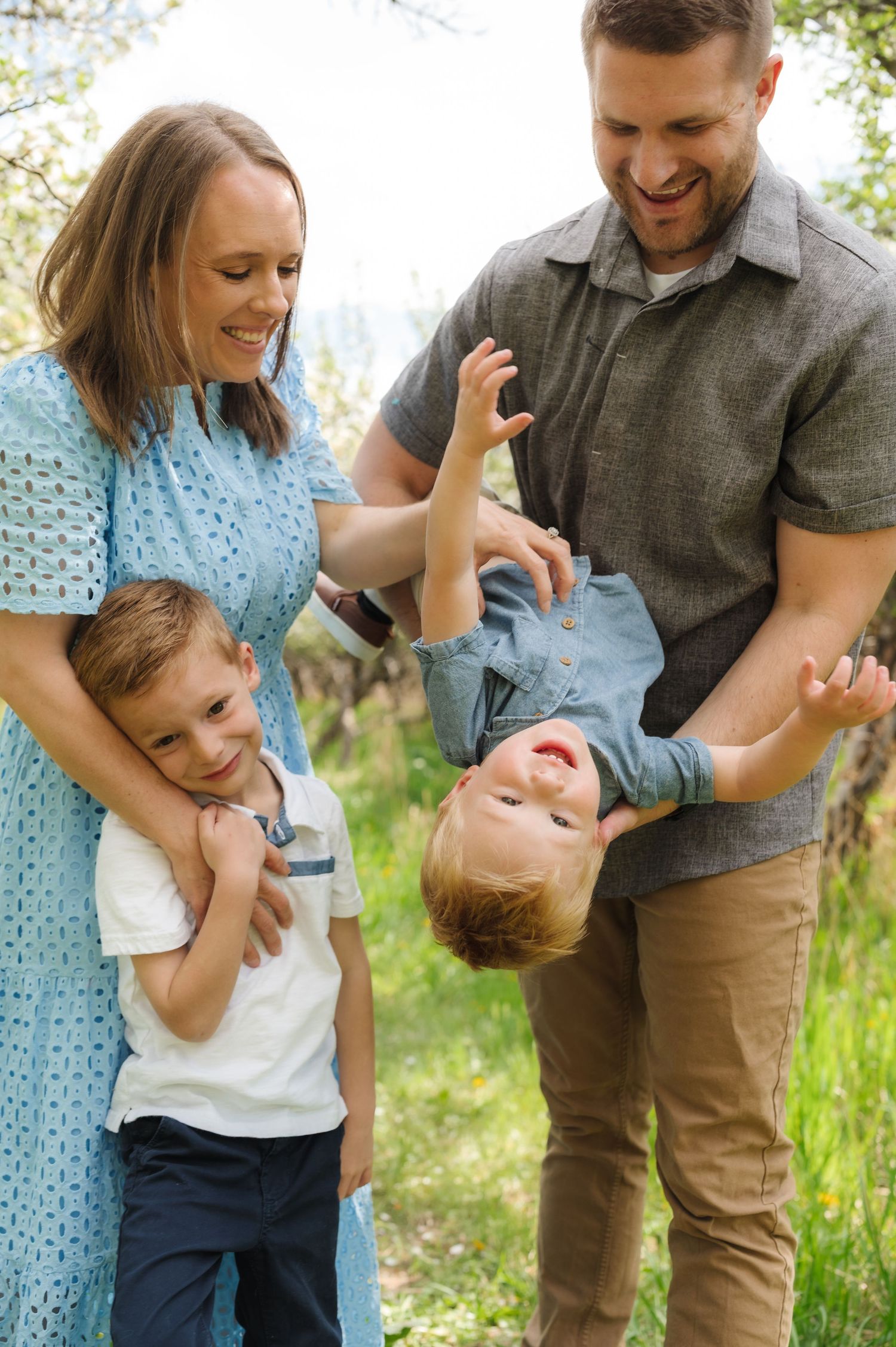 A playful family moment outdoors with a parent tickling a child while another adult watches and laughs in a garden setting.