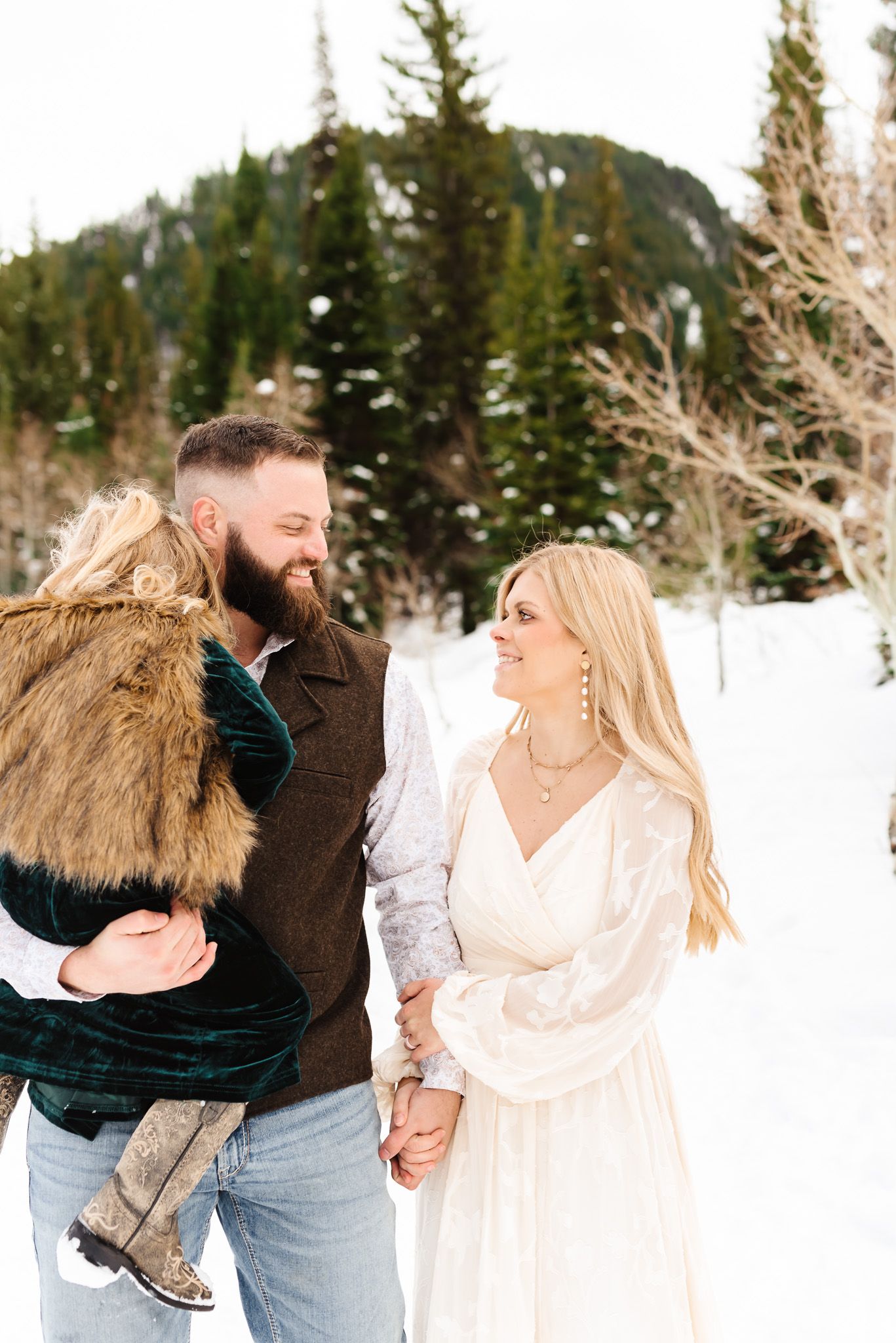 Winter photoshoot in the snow with a couple dressed in cream and brown tones against evergreen trees.