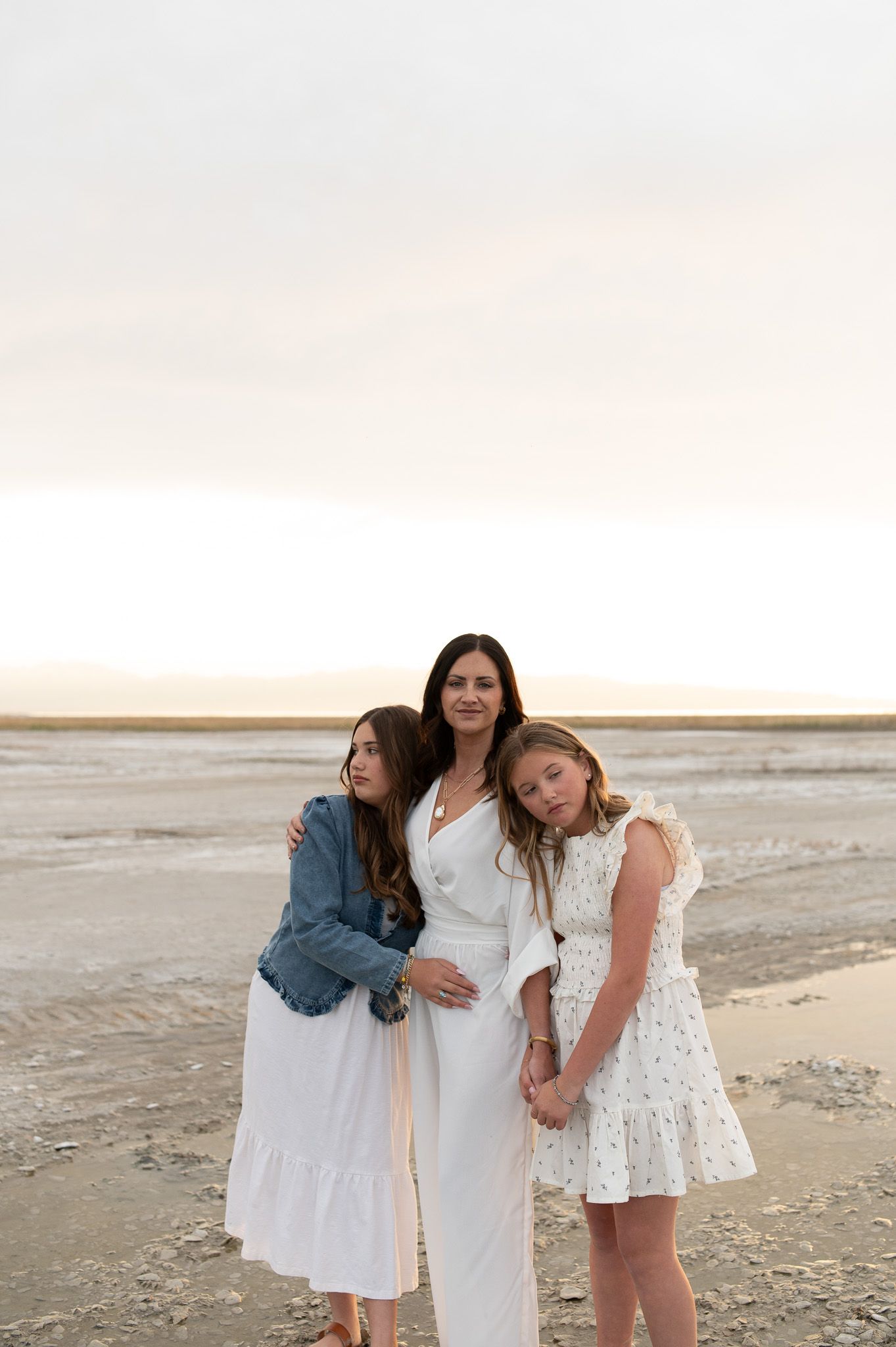 Mother with two daughters posing on beach at sunset in white dresses.