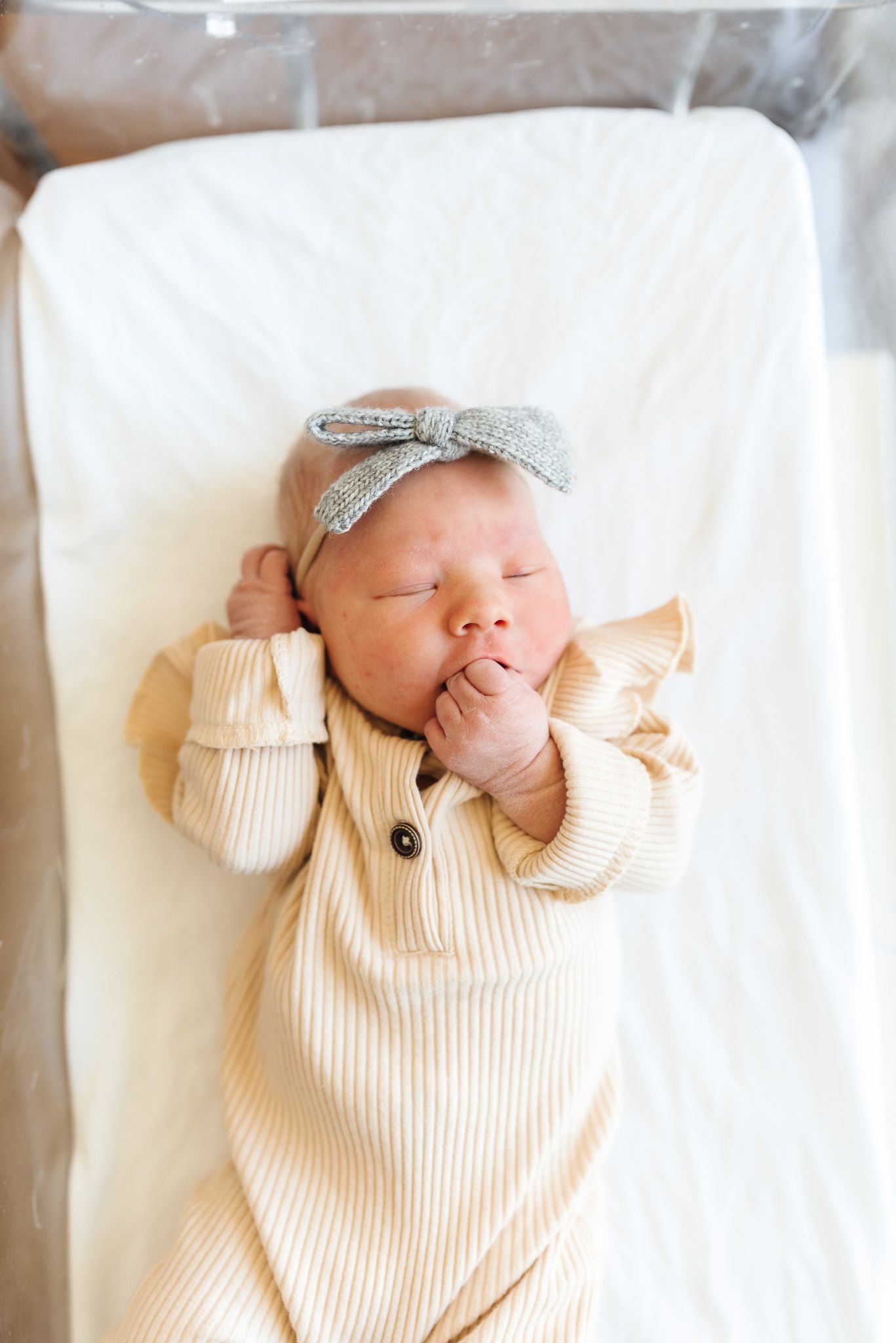 Sleeping newborn baby girl in striped onesie and gray bow headband lying on white hospital bassinet.