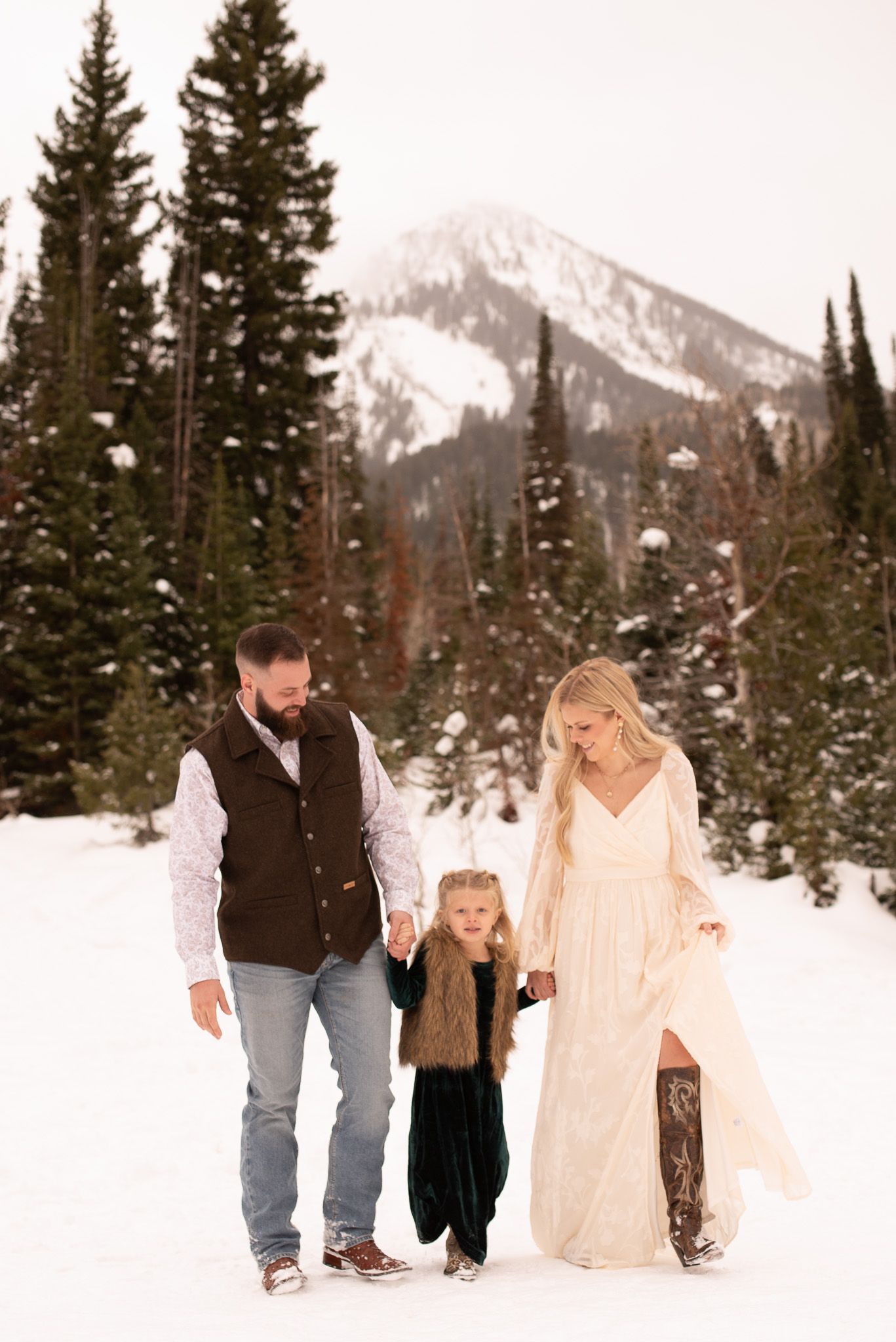 Family of three walks through snowy mountain landscape, bride in white gown and boots, groom in vest, child in fur vest.