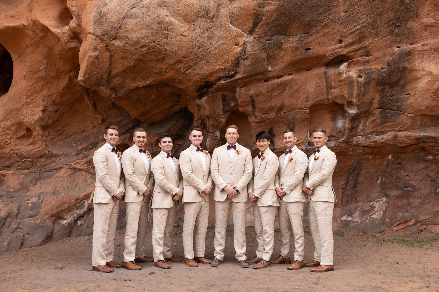 Groom and groomsmen in tan suits posing together in front of red rock formations at an outdoor wedding.