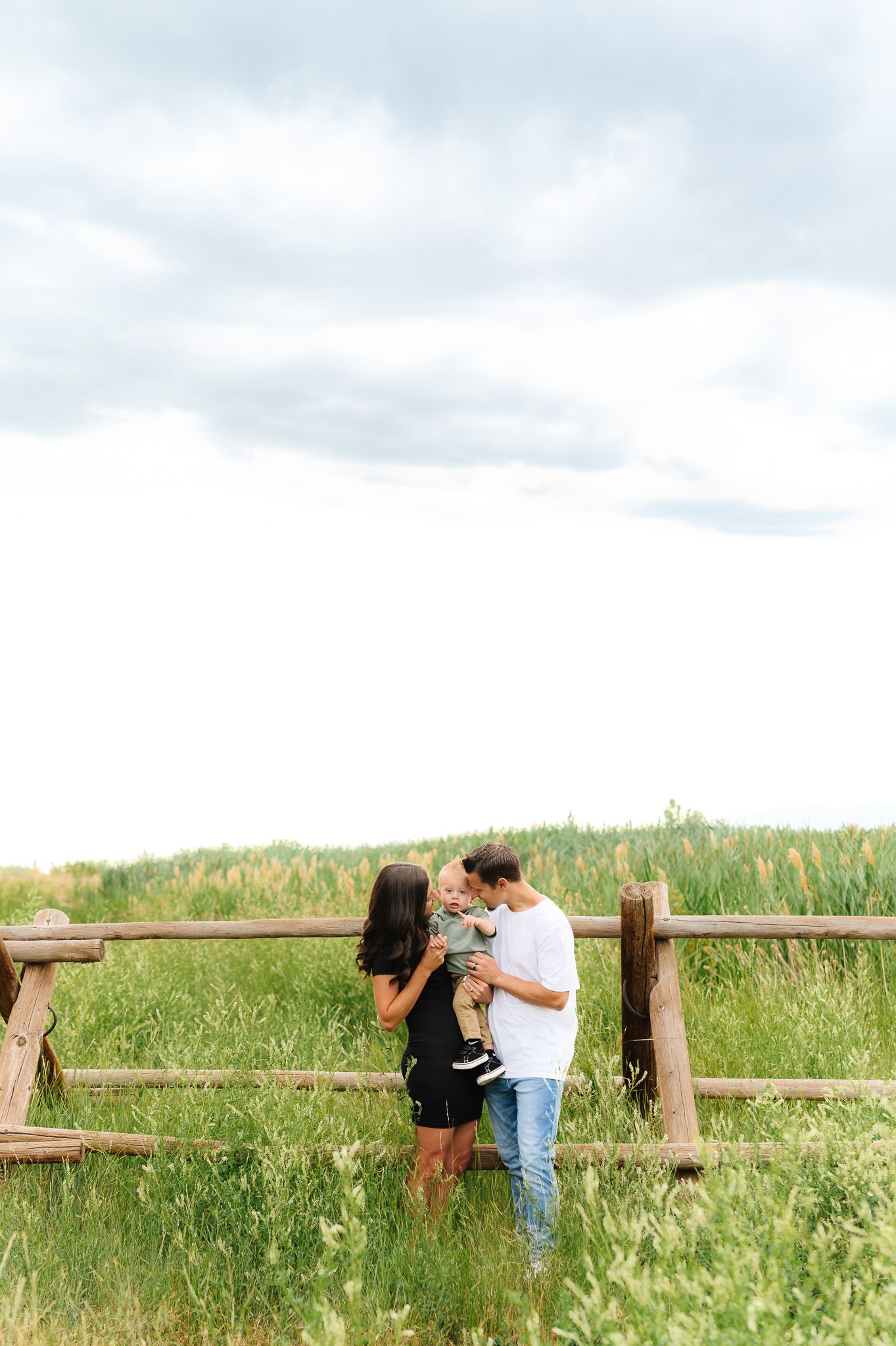 Family enjoying a tender moment together by a wooden fence in a grassy meadow.