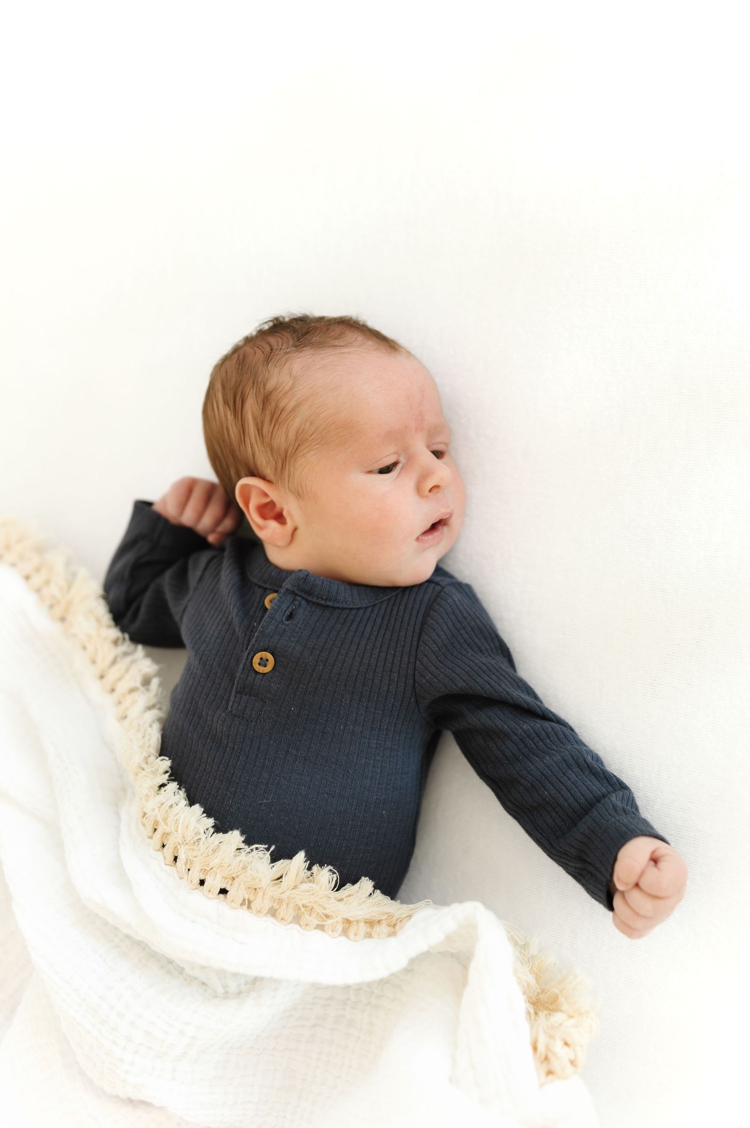Newborn baby in dark ribbed onesie lying on white blanket with fringe trim, looking to the side.
