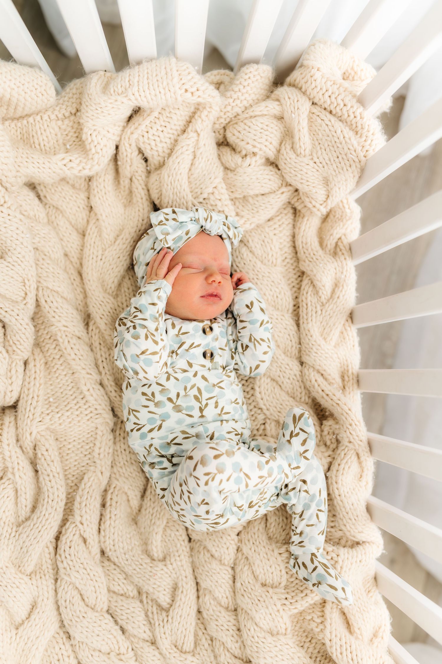 A sleeping newborn wrapped in floral print clothing on a cream knit blanket in a white crib.