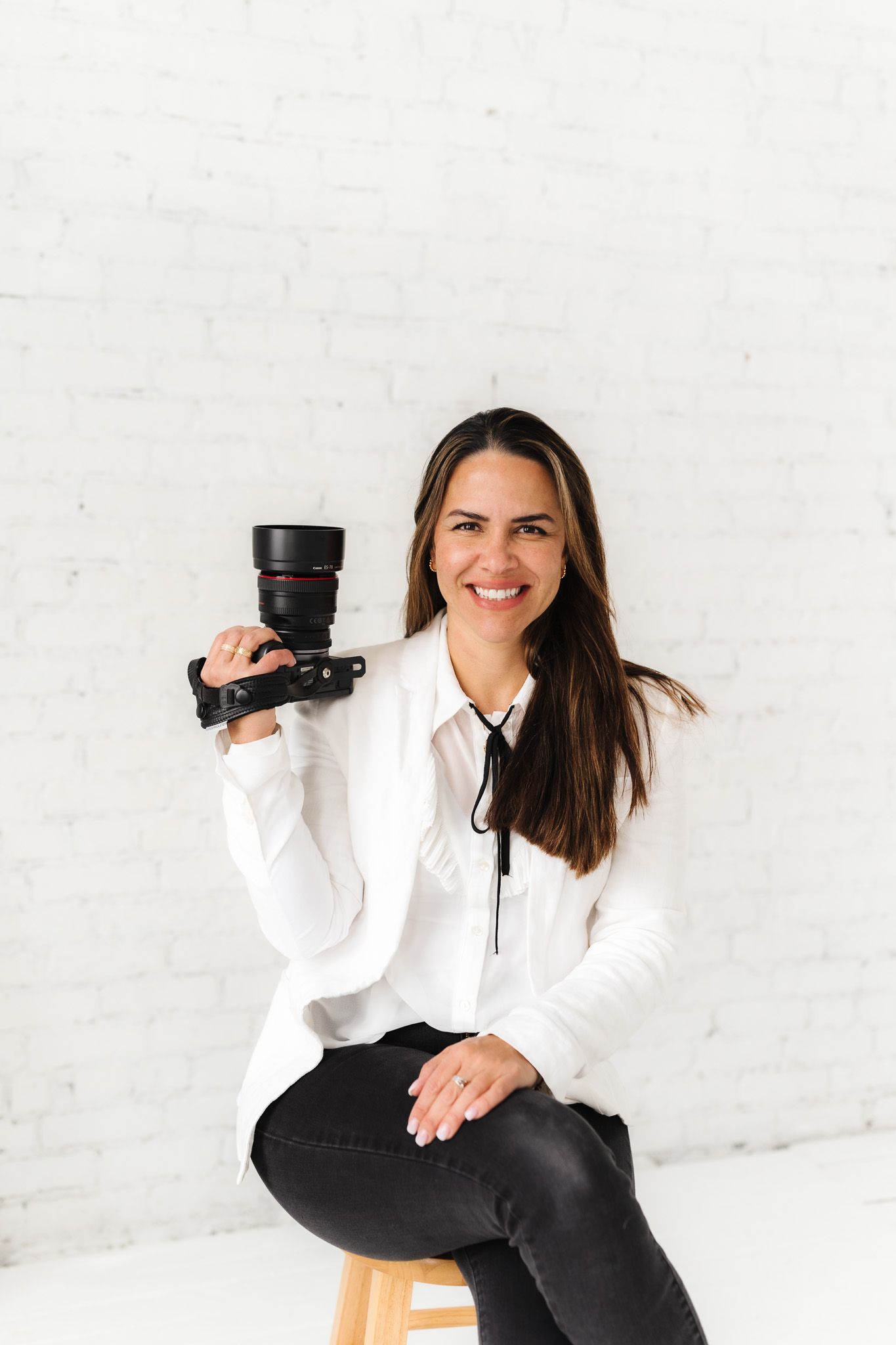 Professional photographer in white blazer holds camera while sitting on wooden stool.