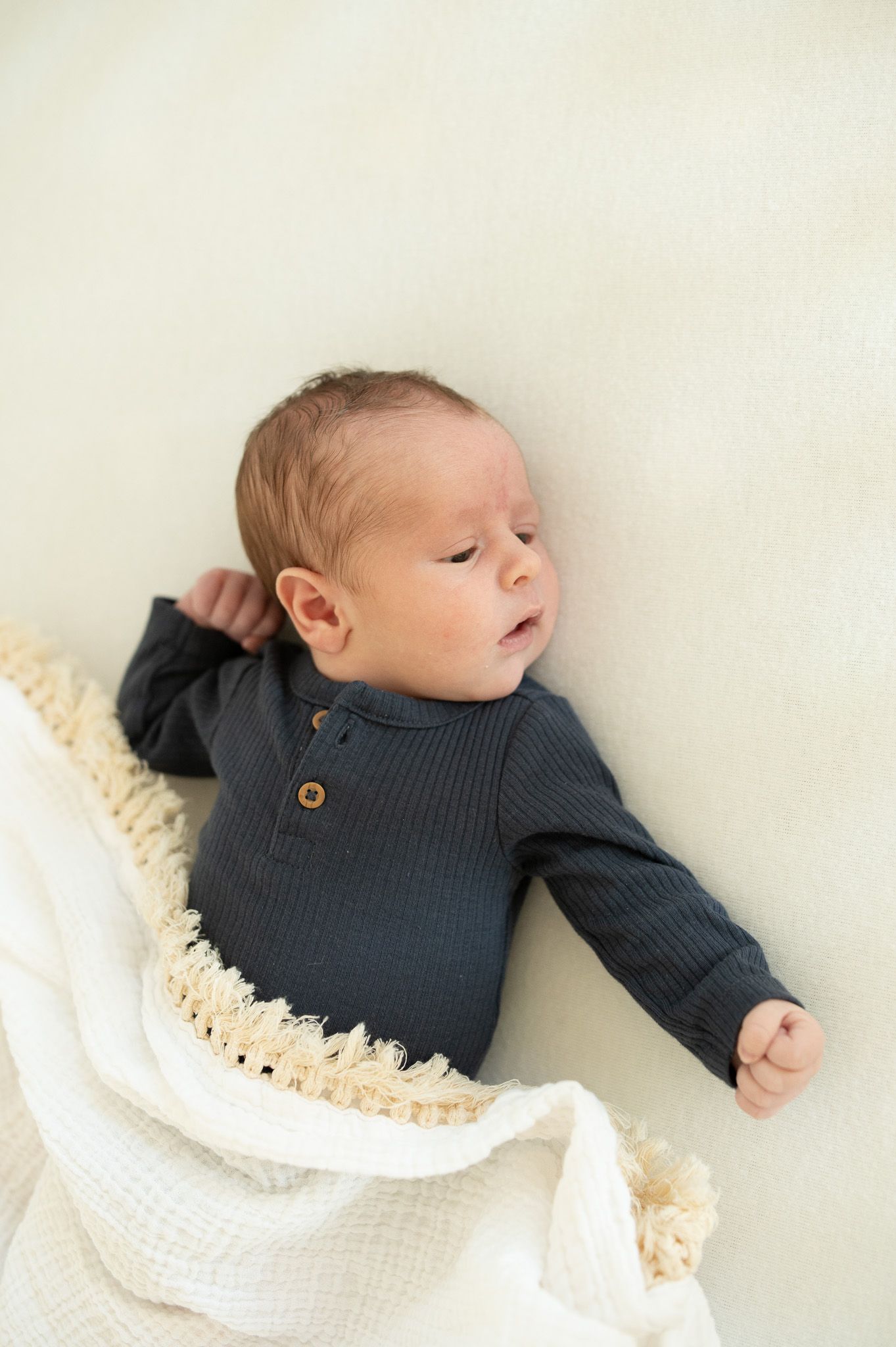 Awake newborn baby in dark gray ribbed onesie with wood buttons lying on cream fringe blanket.