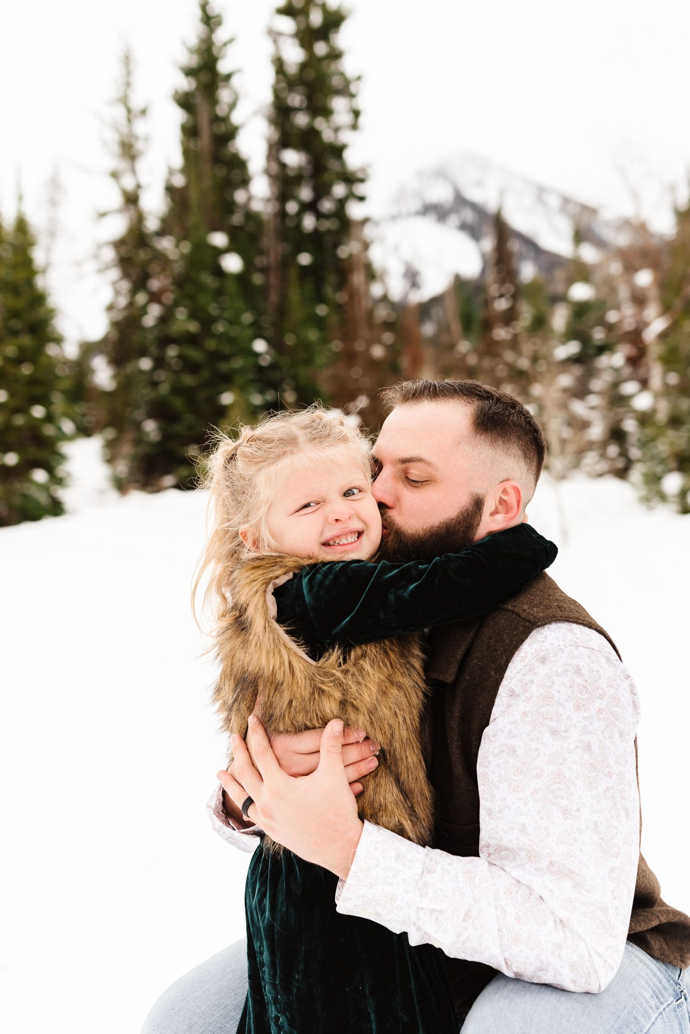 A tender winter photoshoot moment in the snow with evergreen trees in the background.