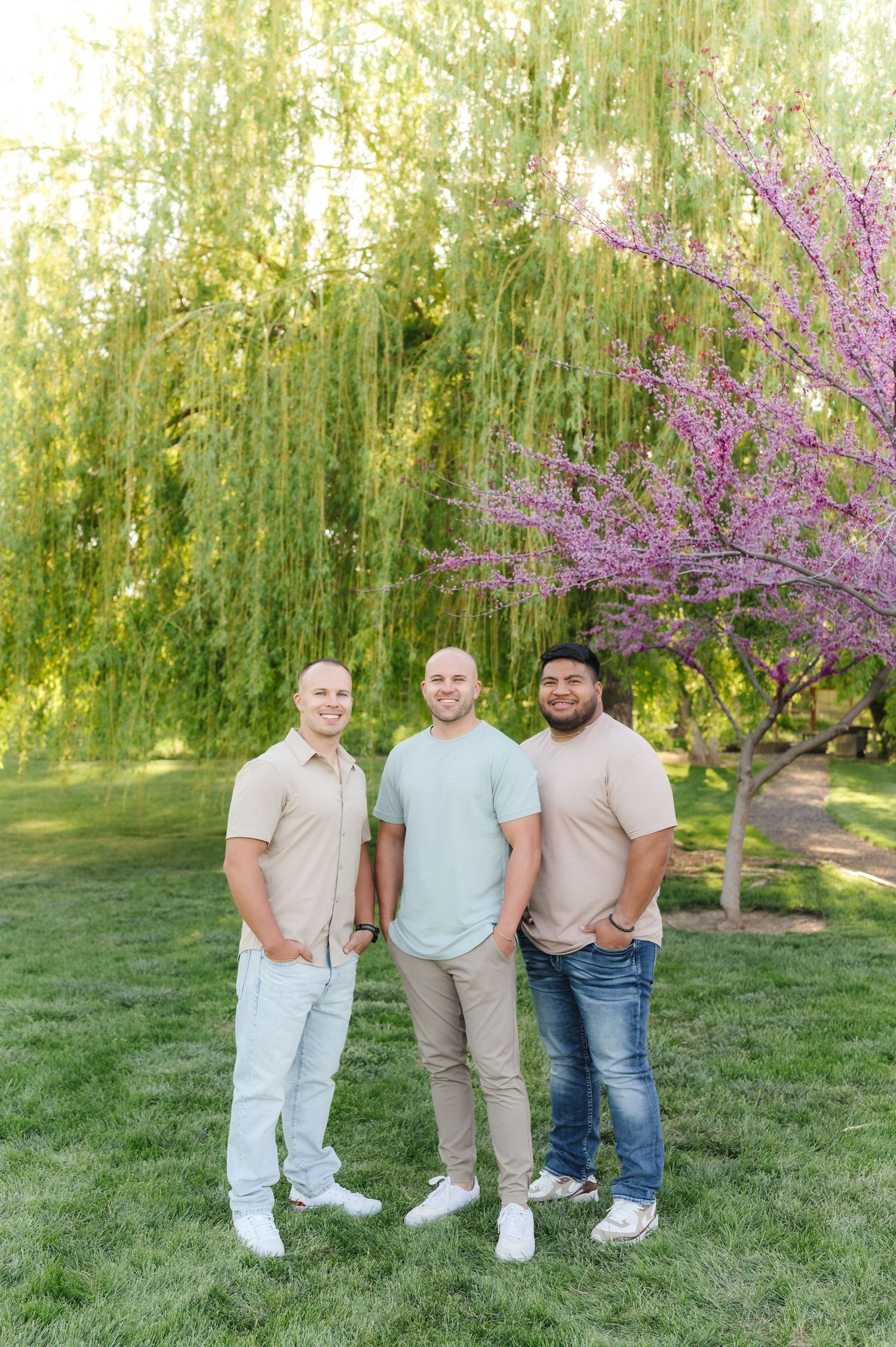 Three people stand together on grass with blooming cherry trees and weeping willows in the background.