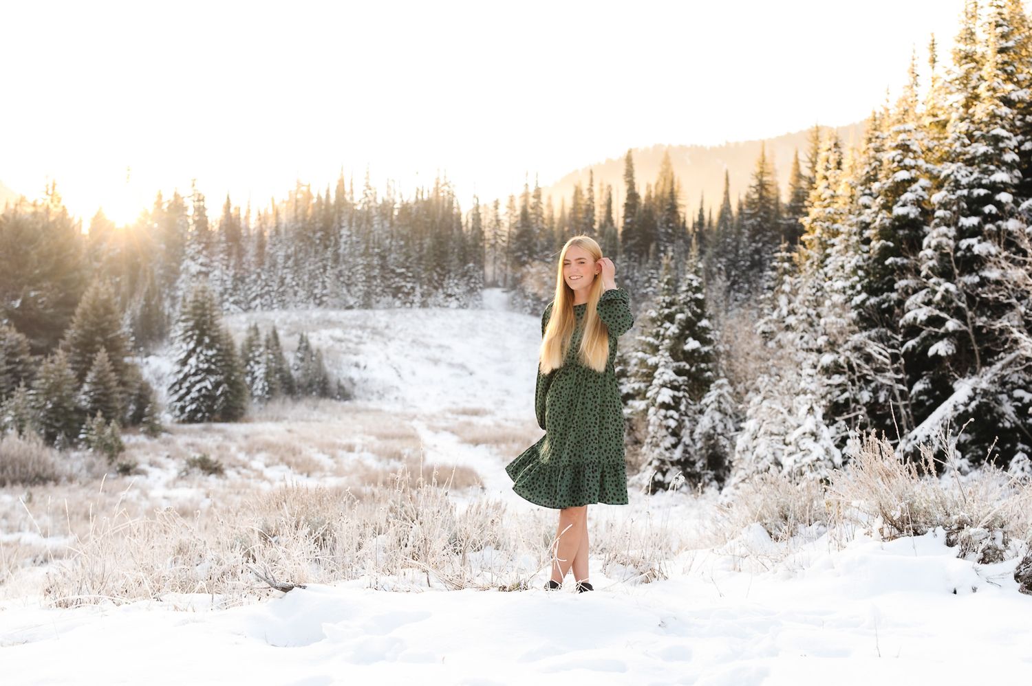 Girl in green dress standing in snowy mountain landscape with snow-covered pine trees and golden sunset light.