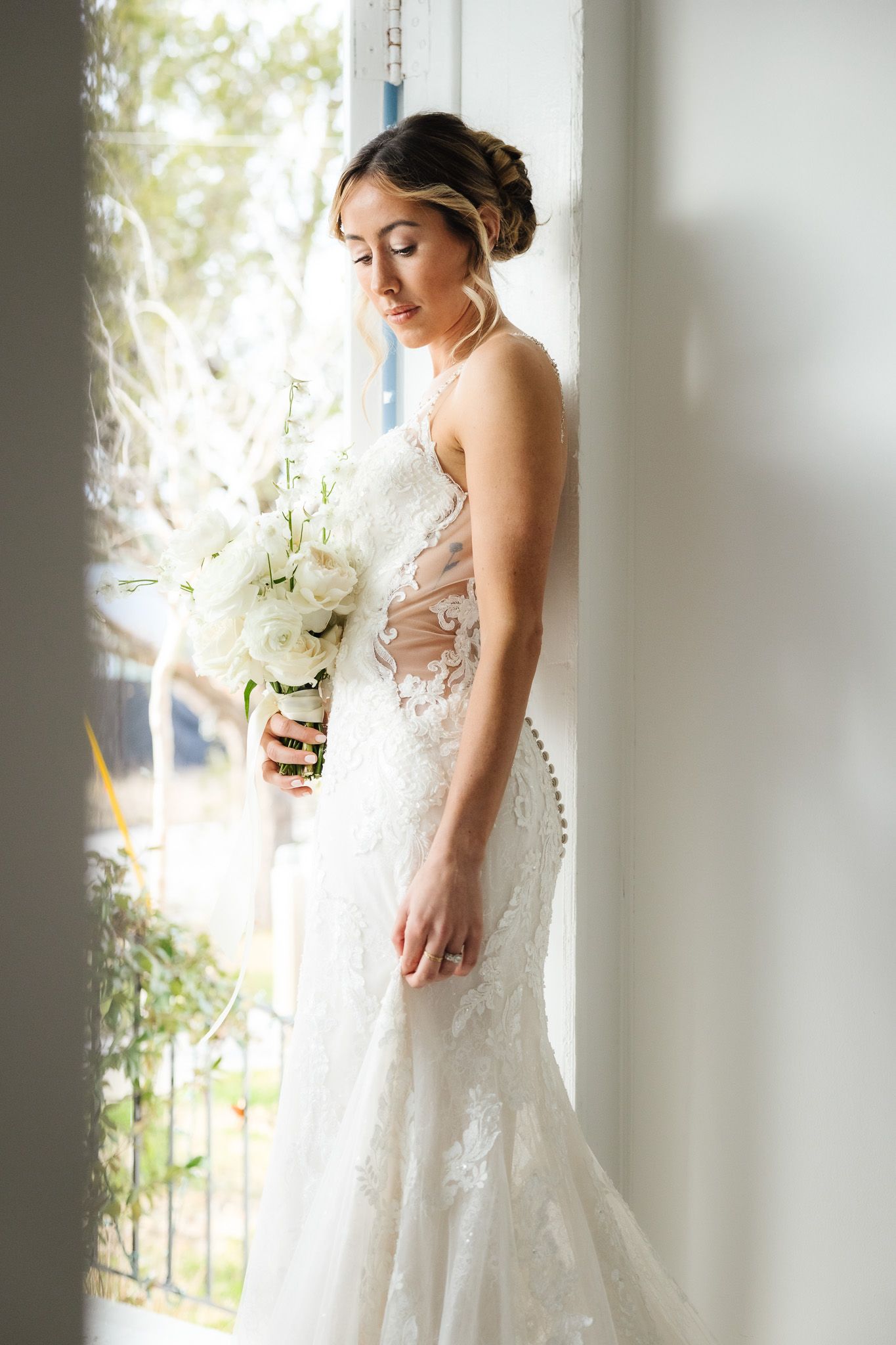 Elegant bride in fitted lace dress poses by bright window with garden view.