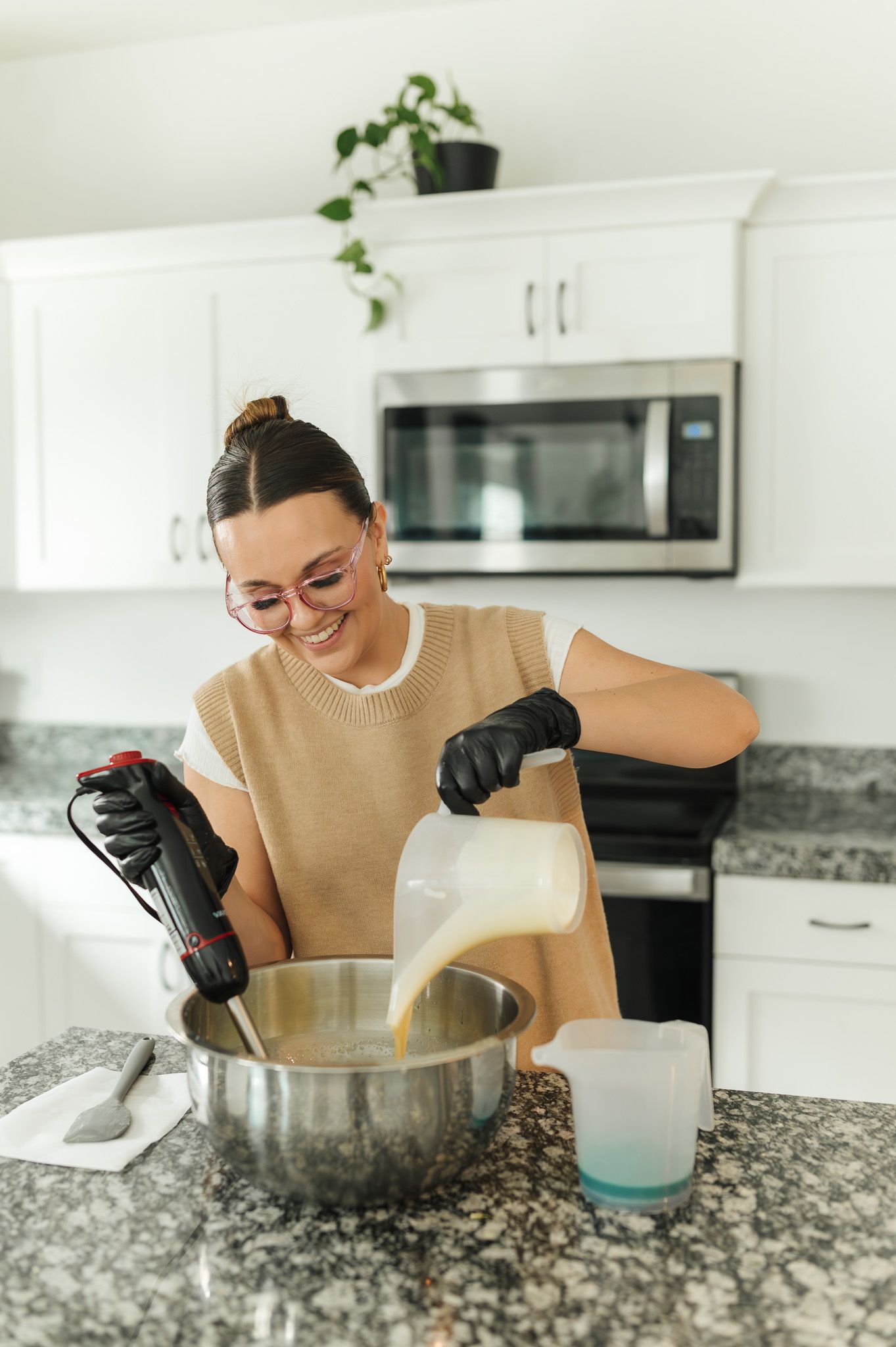 Someone wearing a tan sweater mixes ingredients in a metal bowl on a granite countertop in a modern white kitchen.