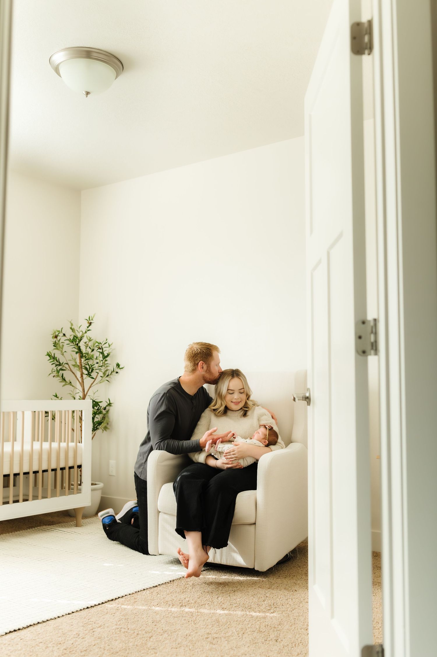 New parents share a tender moment in a nursery, with father kissing mother as she holds their newborn baby in a glider chair.