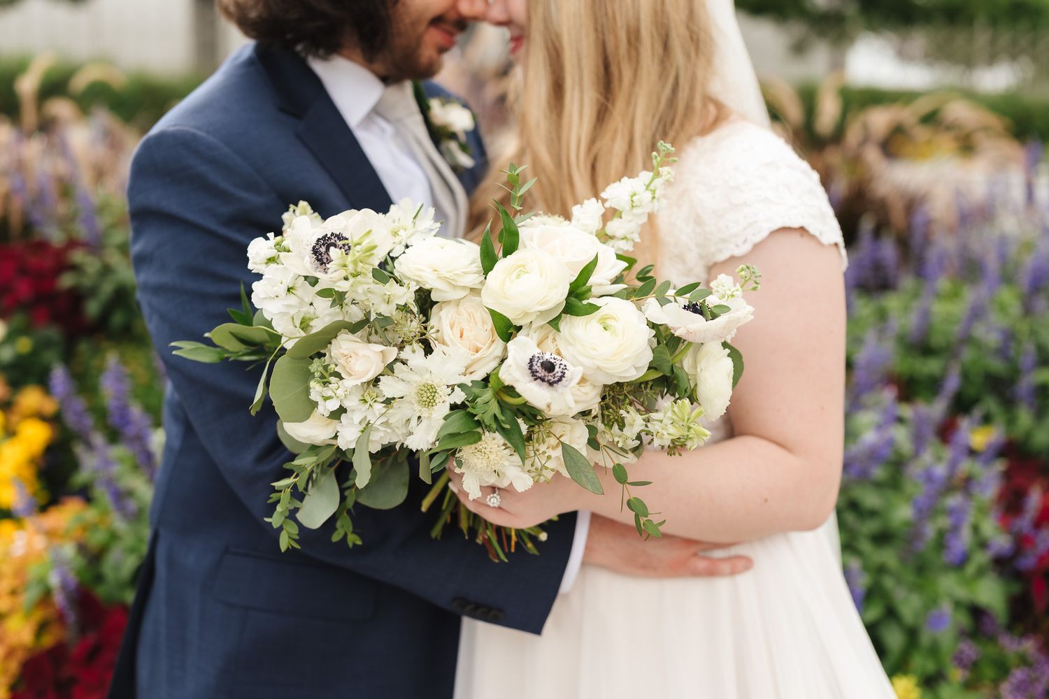 A couple poses with white floral bouquets in a garden setting during their wedding celebration.