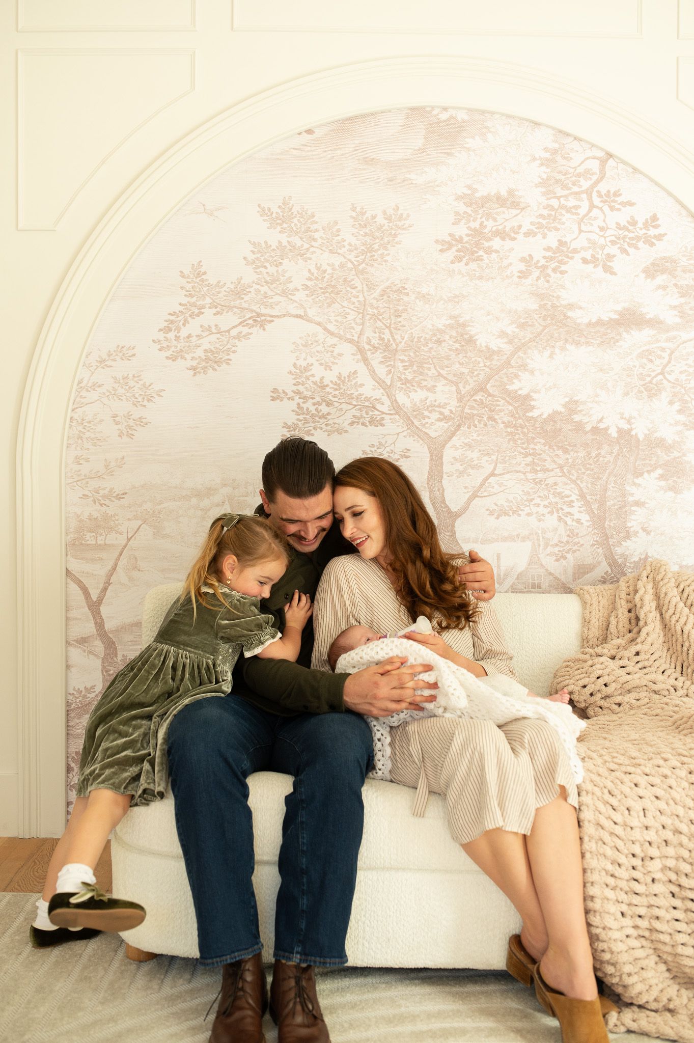Family portrait with parents and toddler girl cuddling newborn baby on white chair before floral mural.