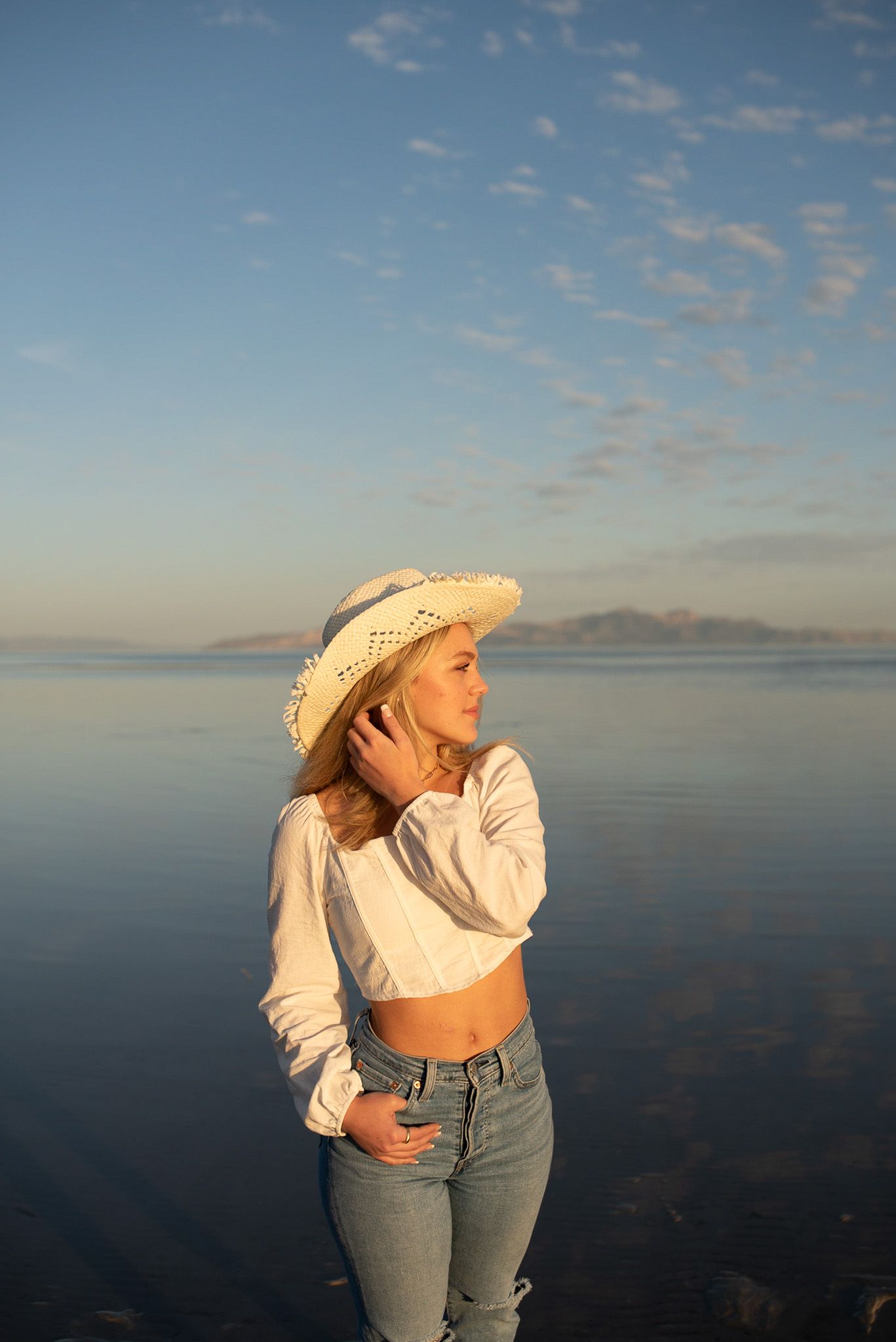 Young blonde woman in white crop top, jeans, and straw cowboy hat poses by calm reflective water at golden hour.