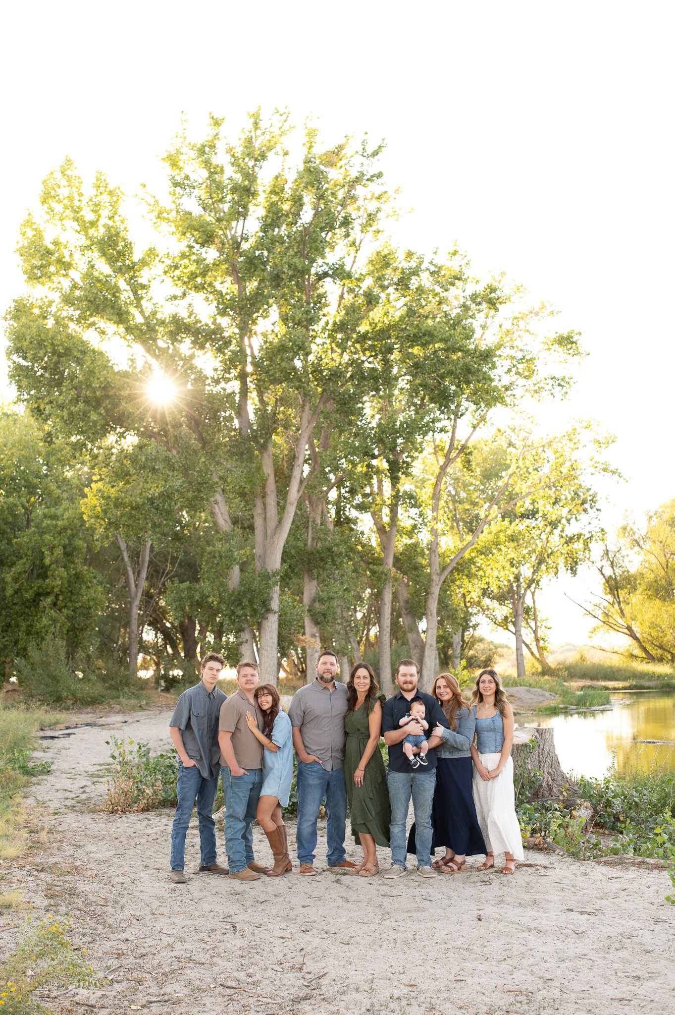 A large family group poses outdoors near a river at golden hour, surrounded by tall green trees on a sandy path.