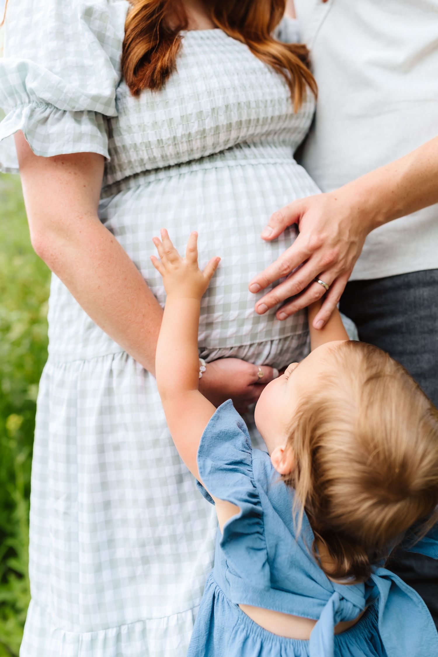 Child in blue dress reaching up to touch a pregnant belly in a gingham dress outdoors.
