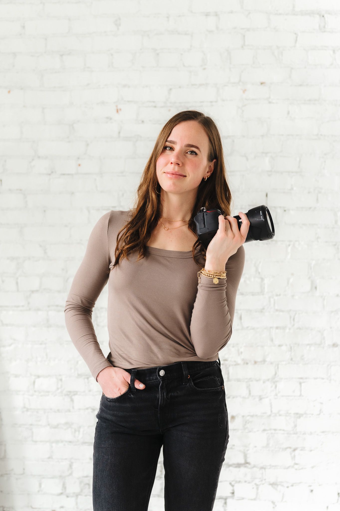 Professional wearing neutral top and dark pants poses with camera in studio setting.