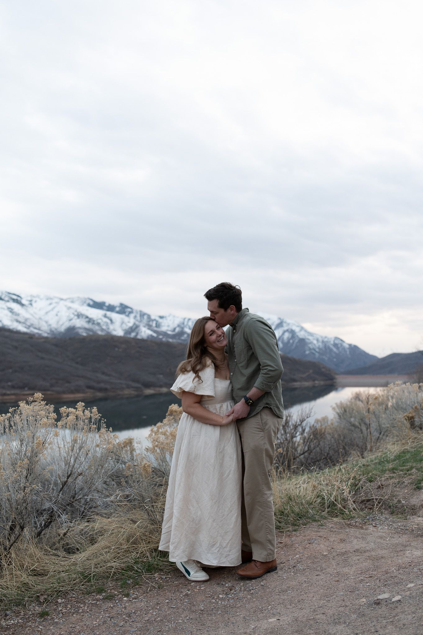 Couple embracing on a scenic overlook with snow-capped mountains and a lake in the background.