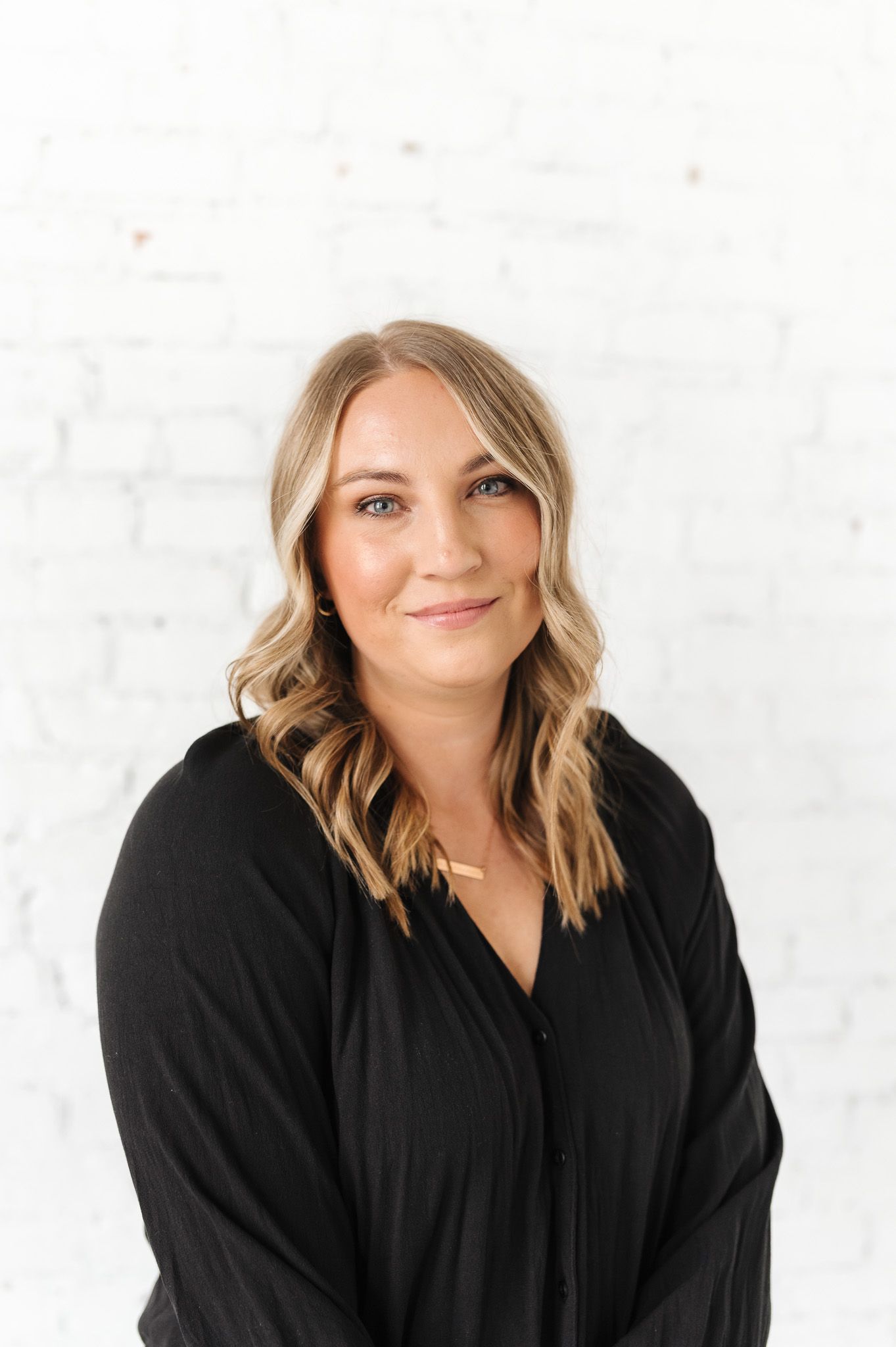 Professional headshot against white brick wall featuring warm smile and wavy hair.