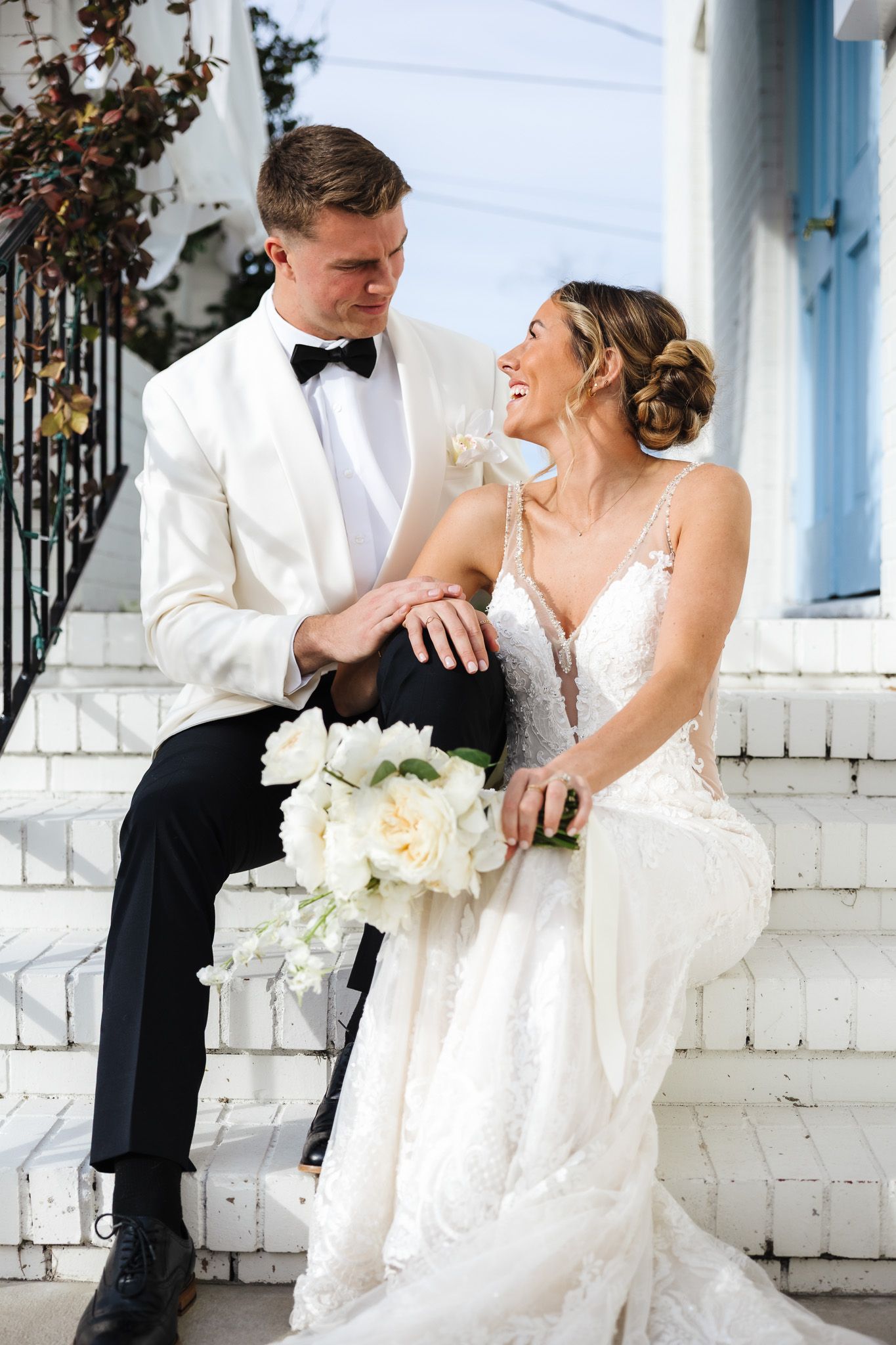 Bride and groom sit together on white brick steps, smiling, as she holds a white peony bouquet in wedding attire.