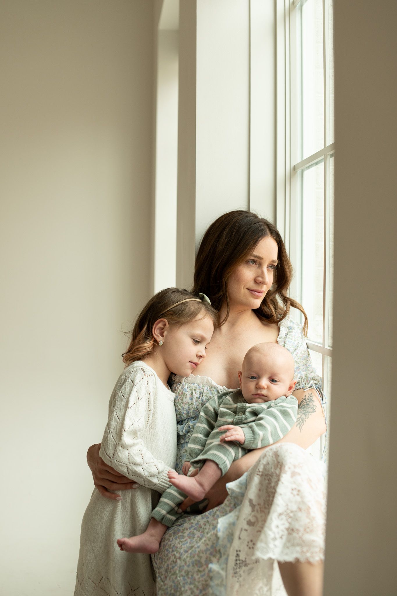 A mother in a floral dress holds her baby and young daughter by a bright window in a serene indoor setting.