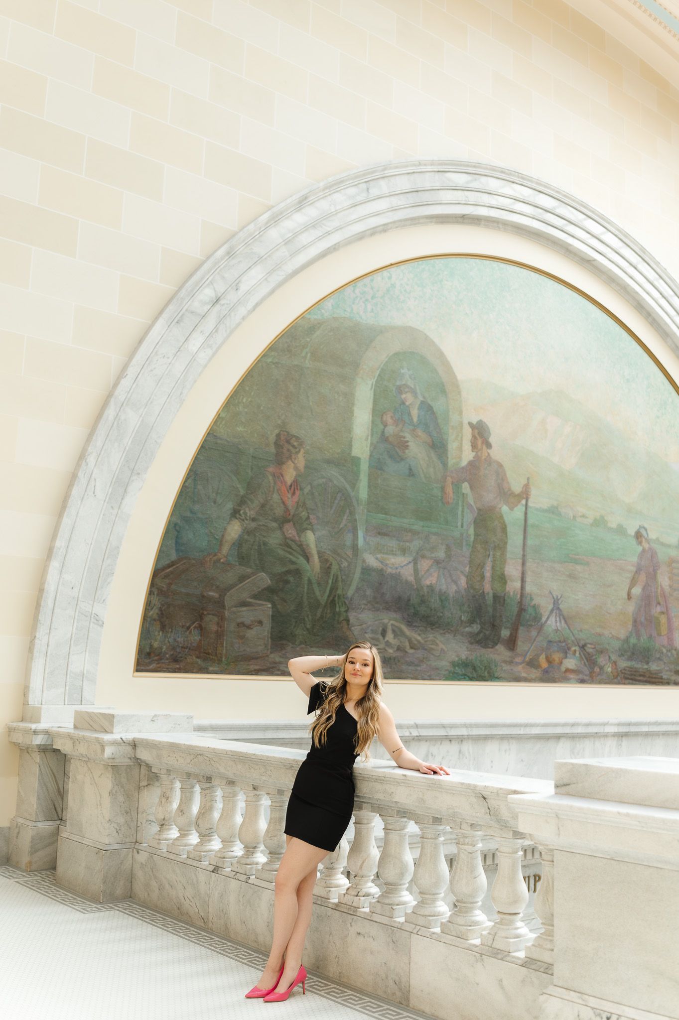 Young woman in black dress and pink heels posing by marble balustrade beneath large classical mural painting.