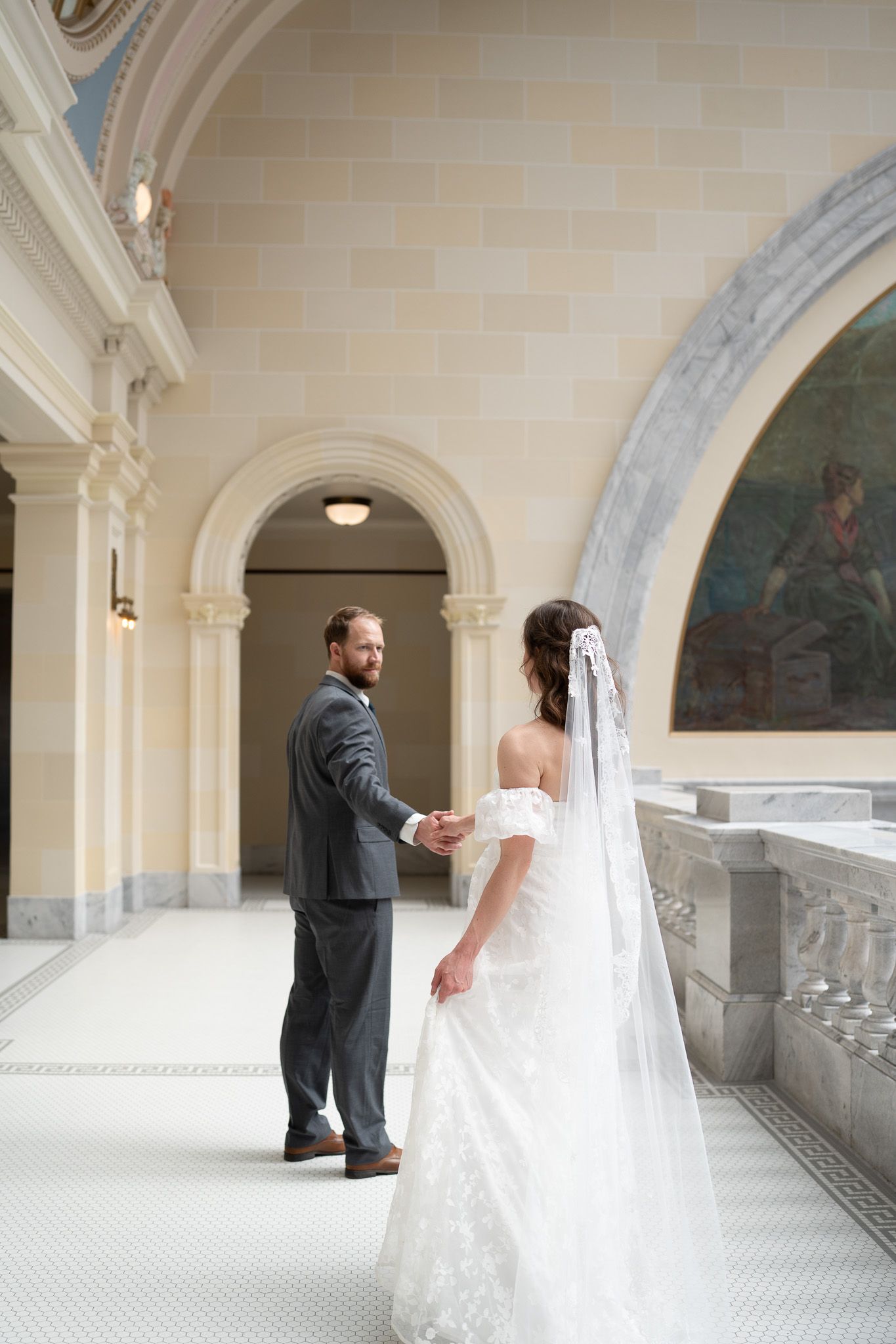 Bride and groom hold hands in elegant columned hall, bride wearing lace gown with cathedral veil.