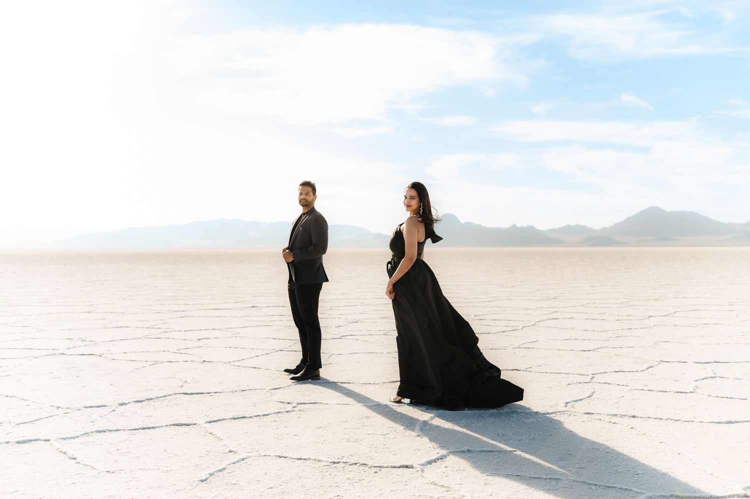 Couple in dark clothing sitting on white salt flats under bright blue sky.