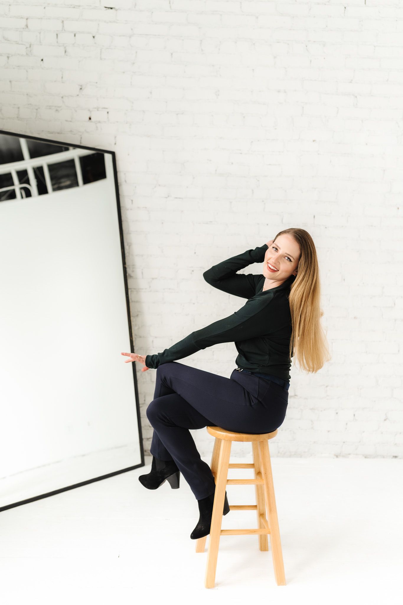 Creative professional in black outfit poses on wooden stool against white backdrop.