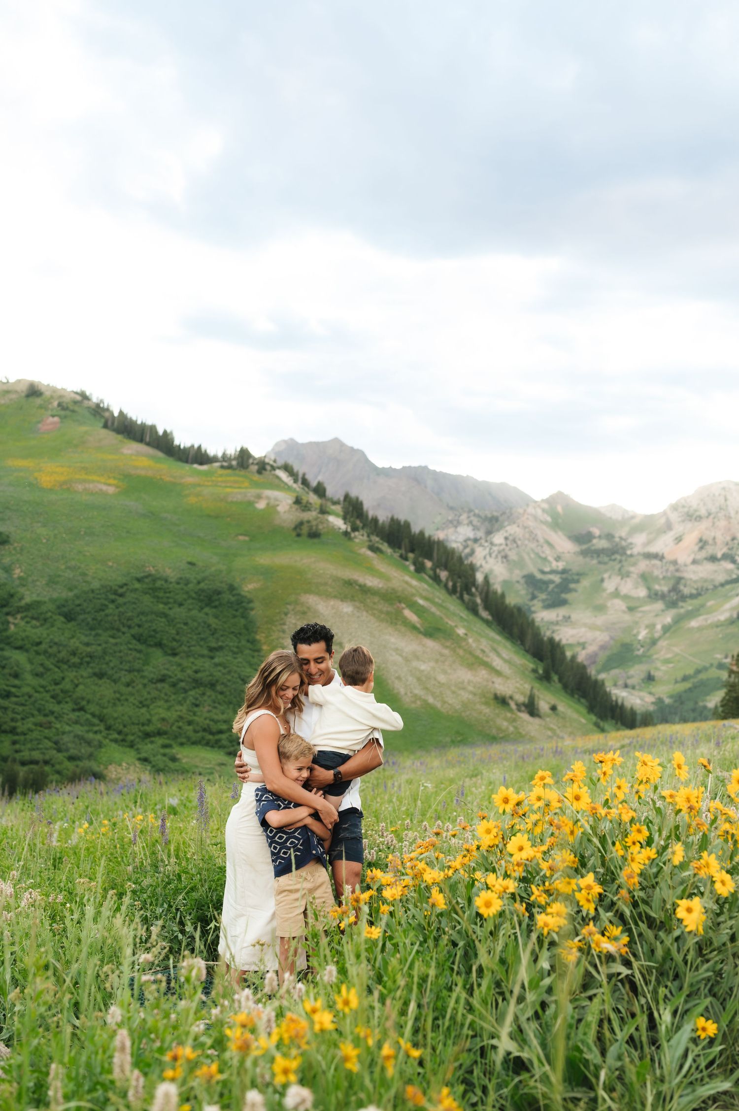 A couple embraces in a mountain meadow filled with vibrant yellow wildflowers and rolling hills in the background.