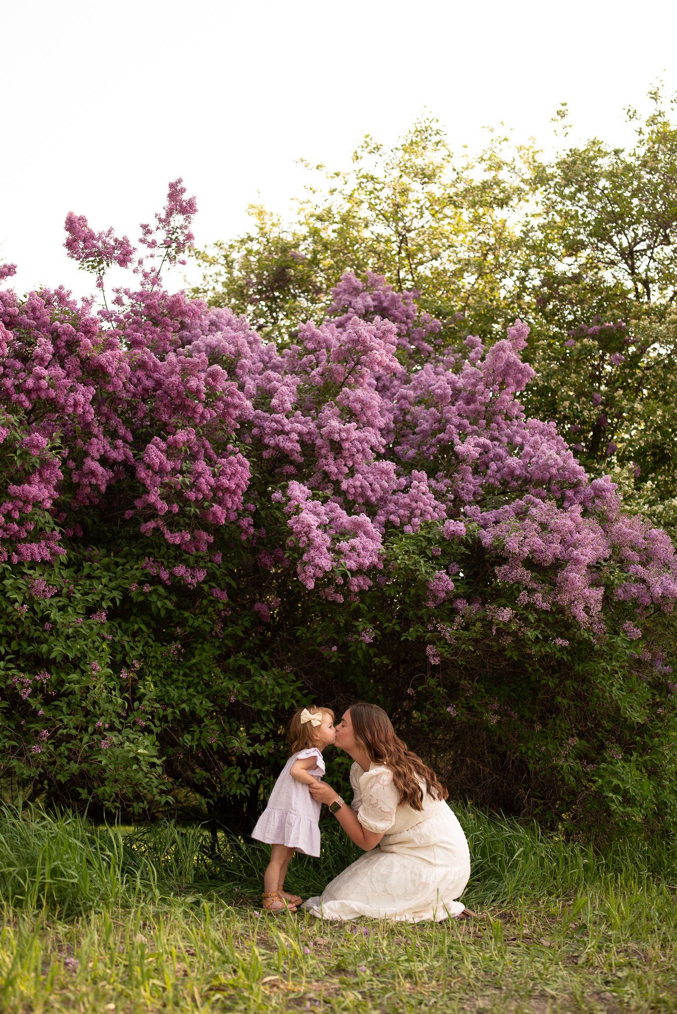 Mother kissing toddler daughter in front of blooming purple lilac bushes outdoors.