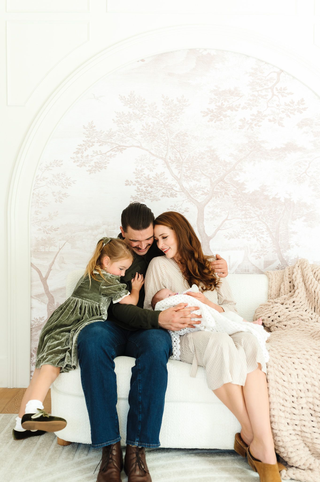 A family of four sits together on a white chair, parents gazing lovingly at their newborn baby while their toddler daughter leans in.