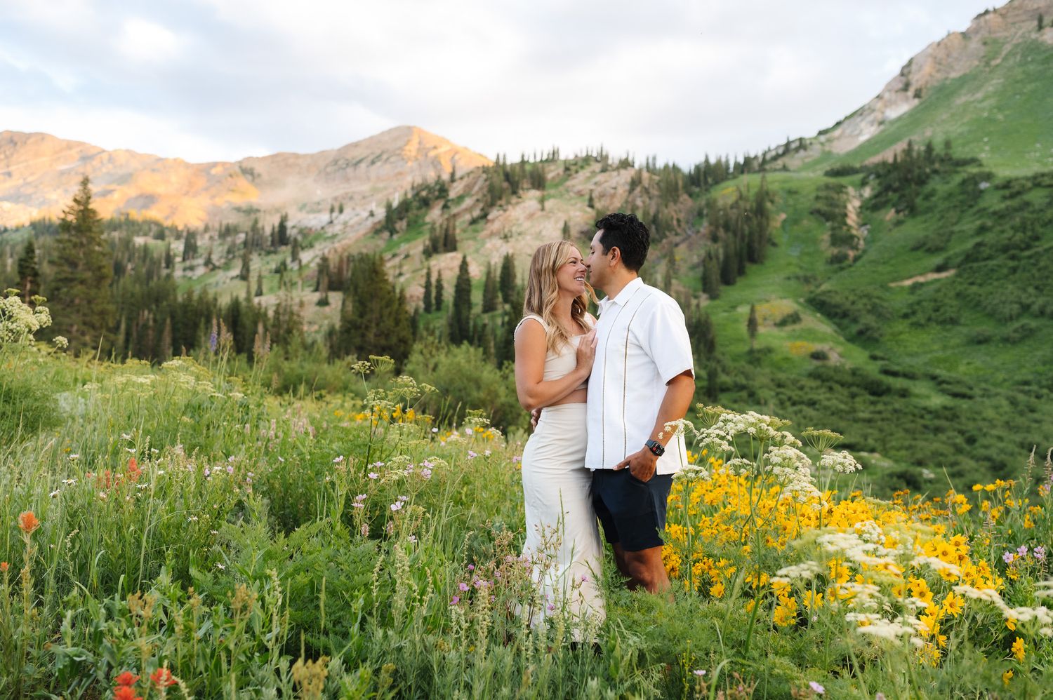 Couple embraces in a mountain meadow filled with vibrant yellow wildflowers and dramatic peaks in the background.