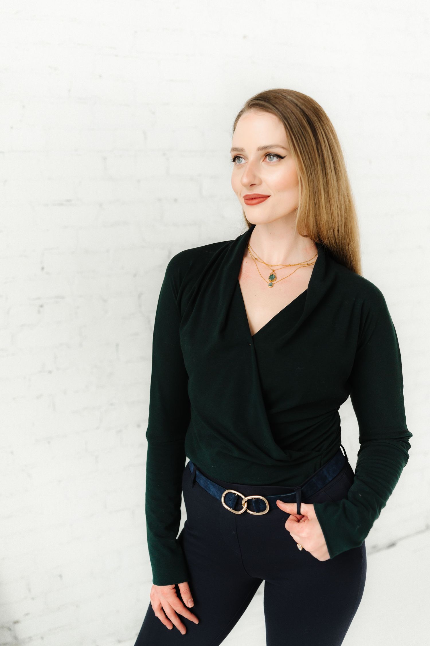 Woman in black wrap dress posing against white wall showing jewelry styling.