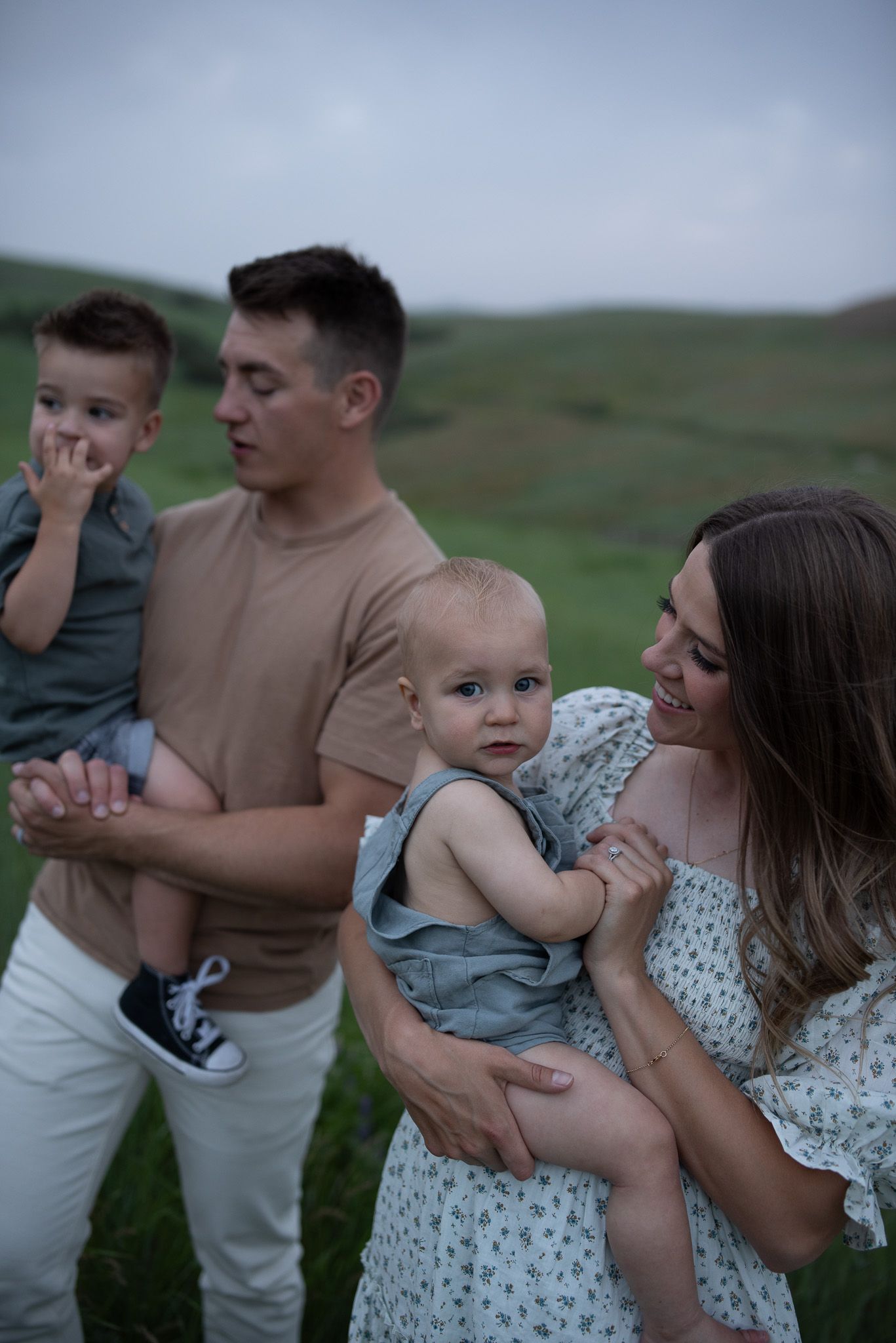 A young family with two small children poses outdoors on a green hillside on a cloudy day.