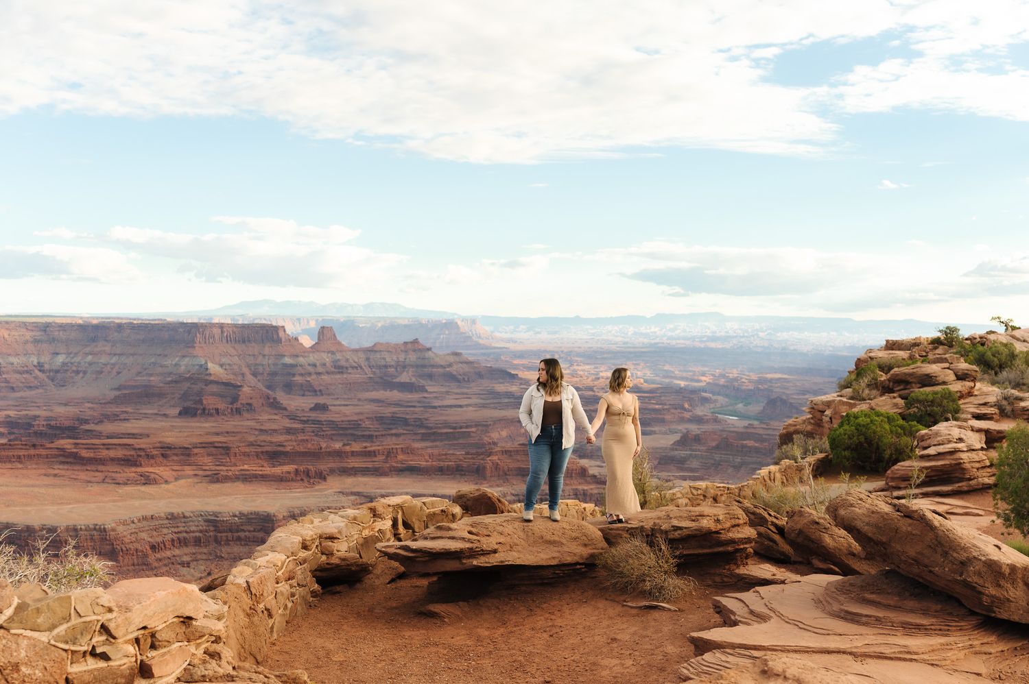 Couple holding hands on rocky cliff overlooking vast canyon landscape in Moab Utah.