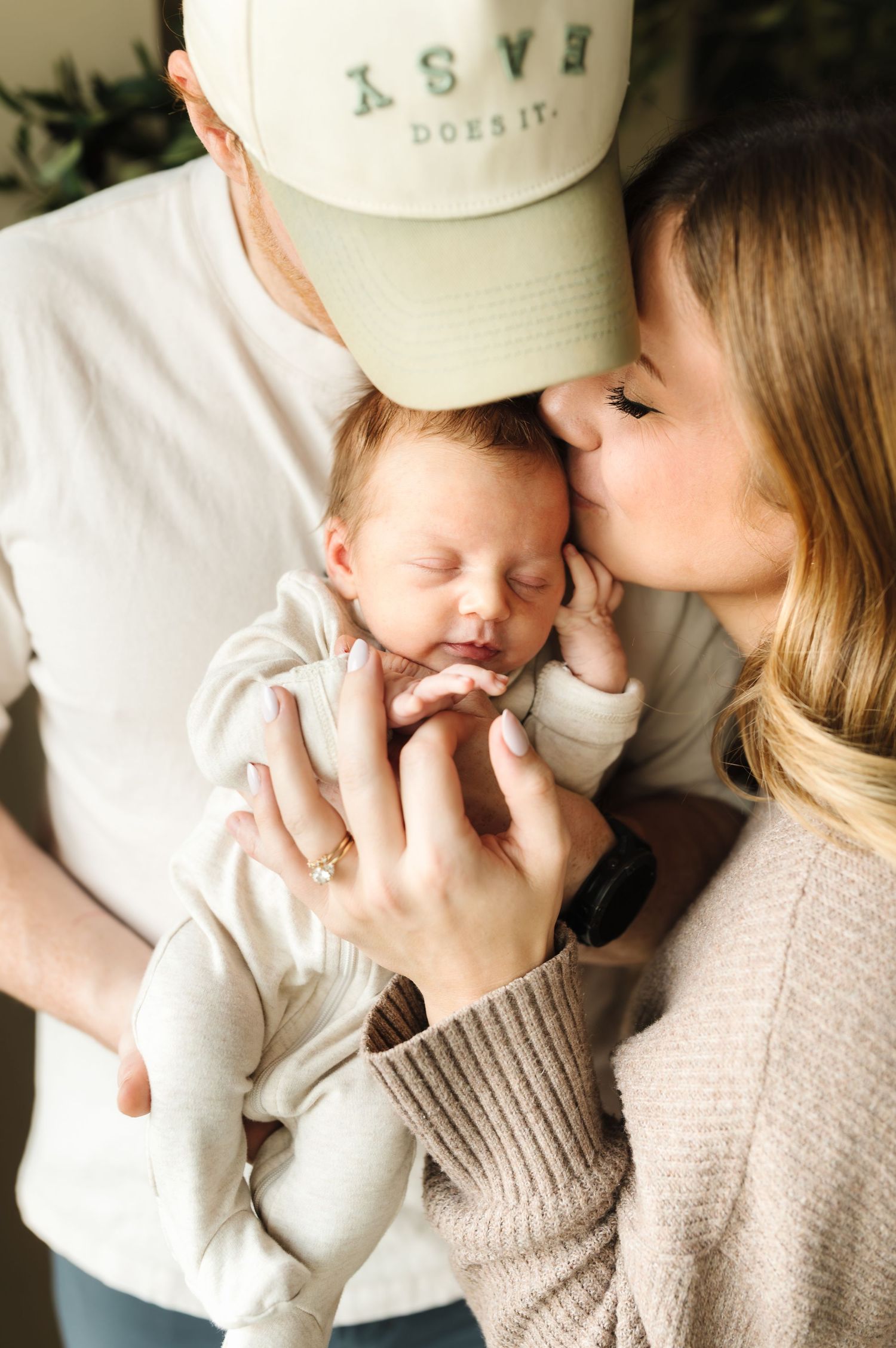 New parents tenderly kiss their sleeping newborn baby, mother wearing a beige sweater and father in a white cap.