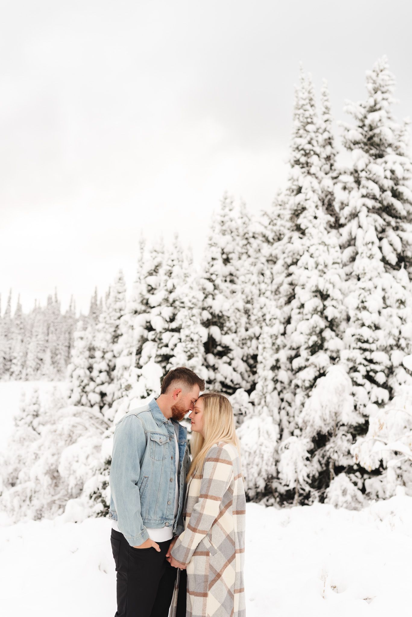 Couple sharing a romantic moment in a snowy winter landscape with snow-covered evergreen trees behind them.