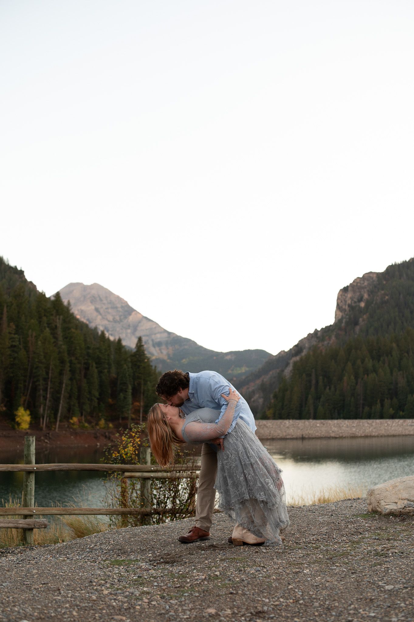 Couple sharing a romantic dip kiss by a mountain lake with pine trees and peaks in the background.