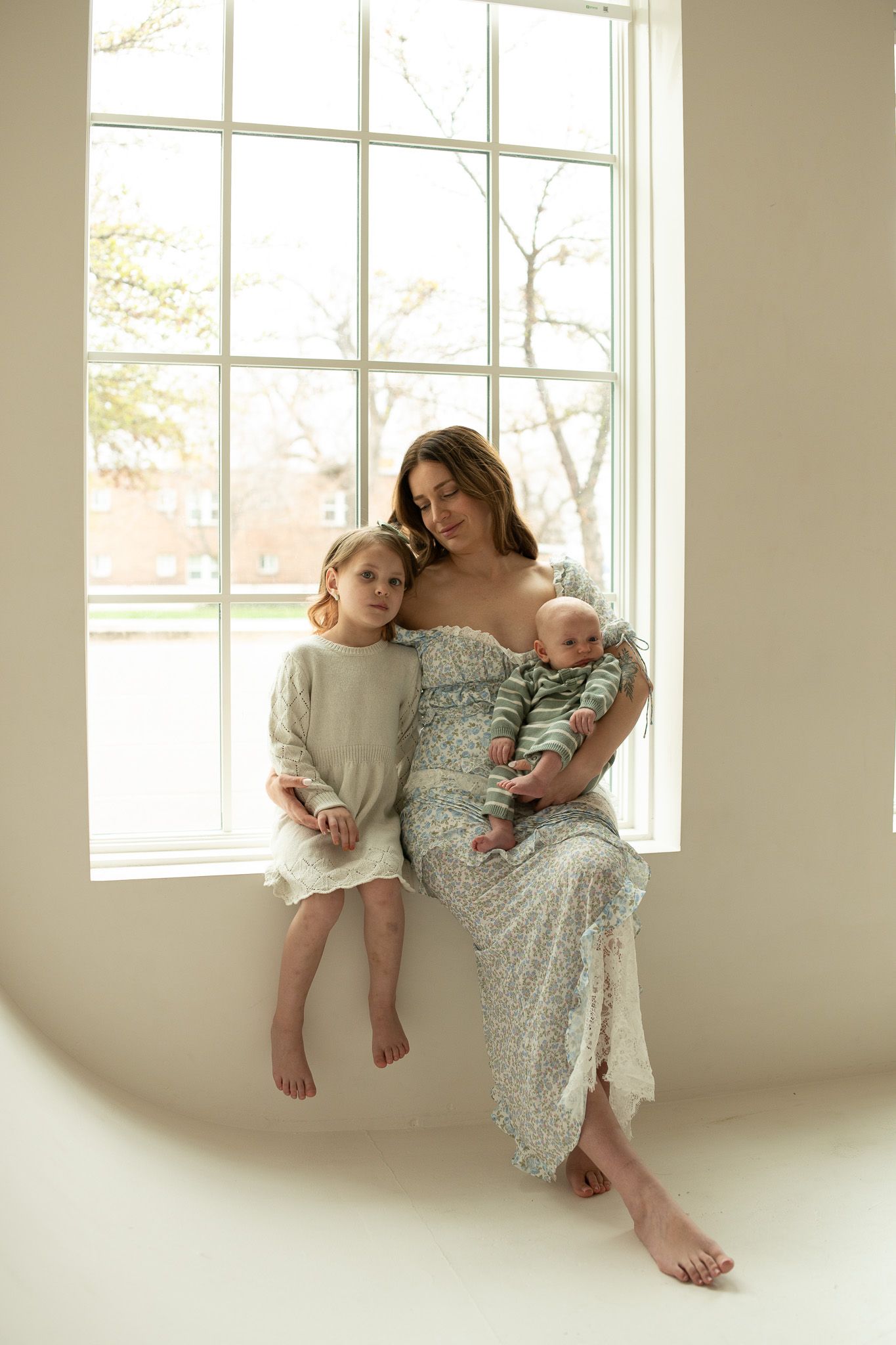 Mother and children in matching floral dresses pose by bright window in minimalist setting.