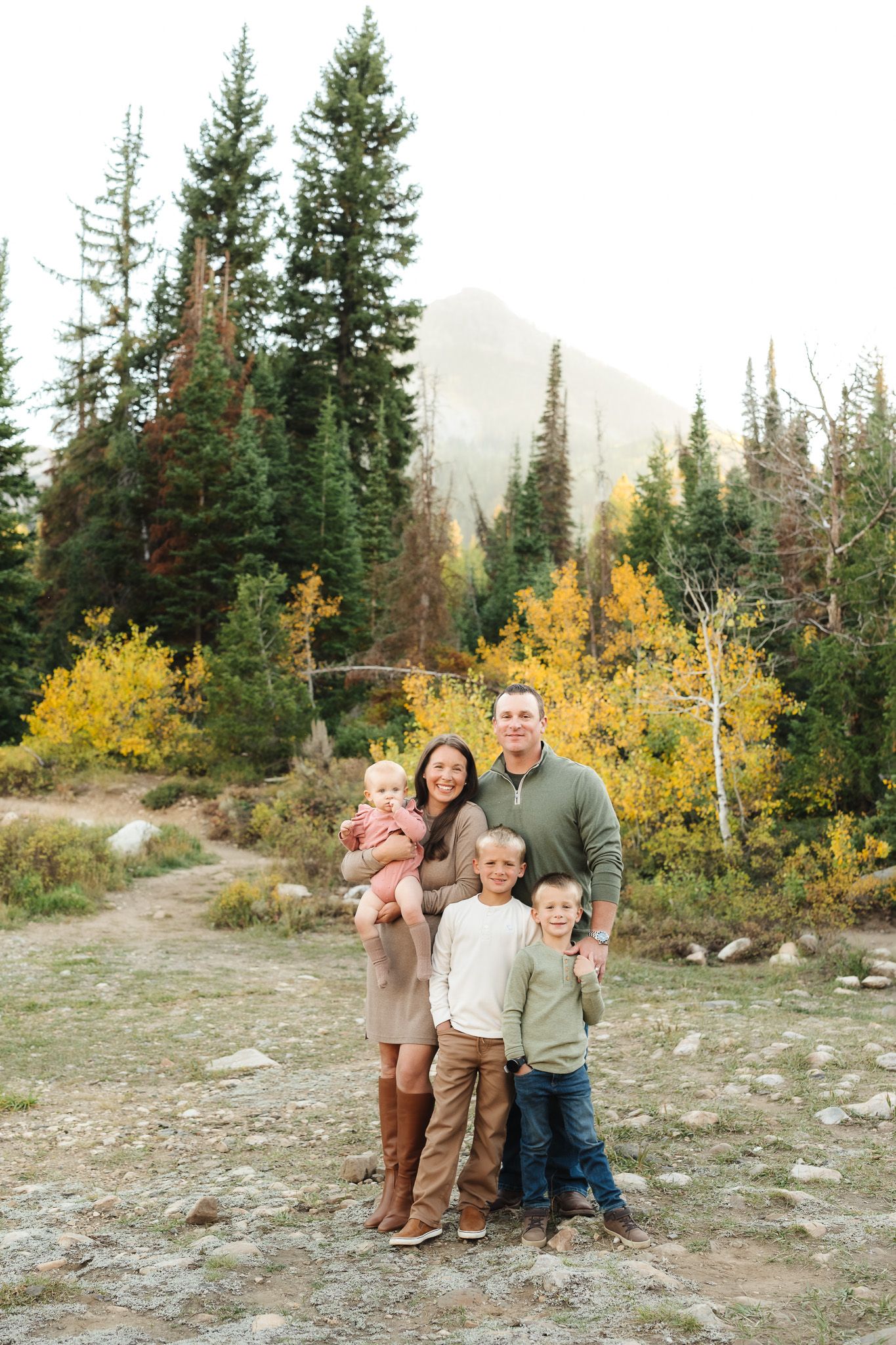 Family portrait in casual attire standing together on rocky terrain with mountain and forest backdrop.