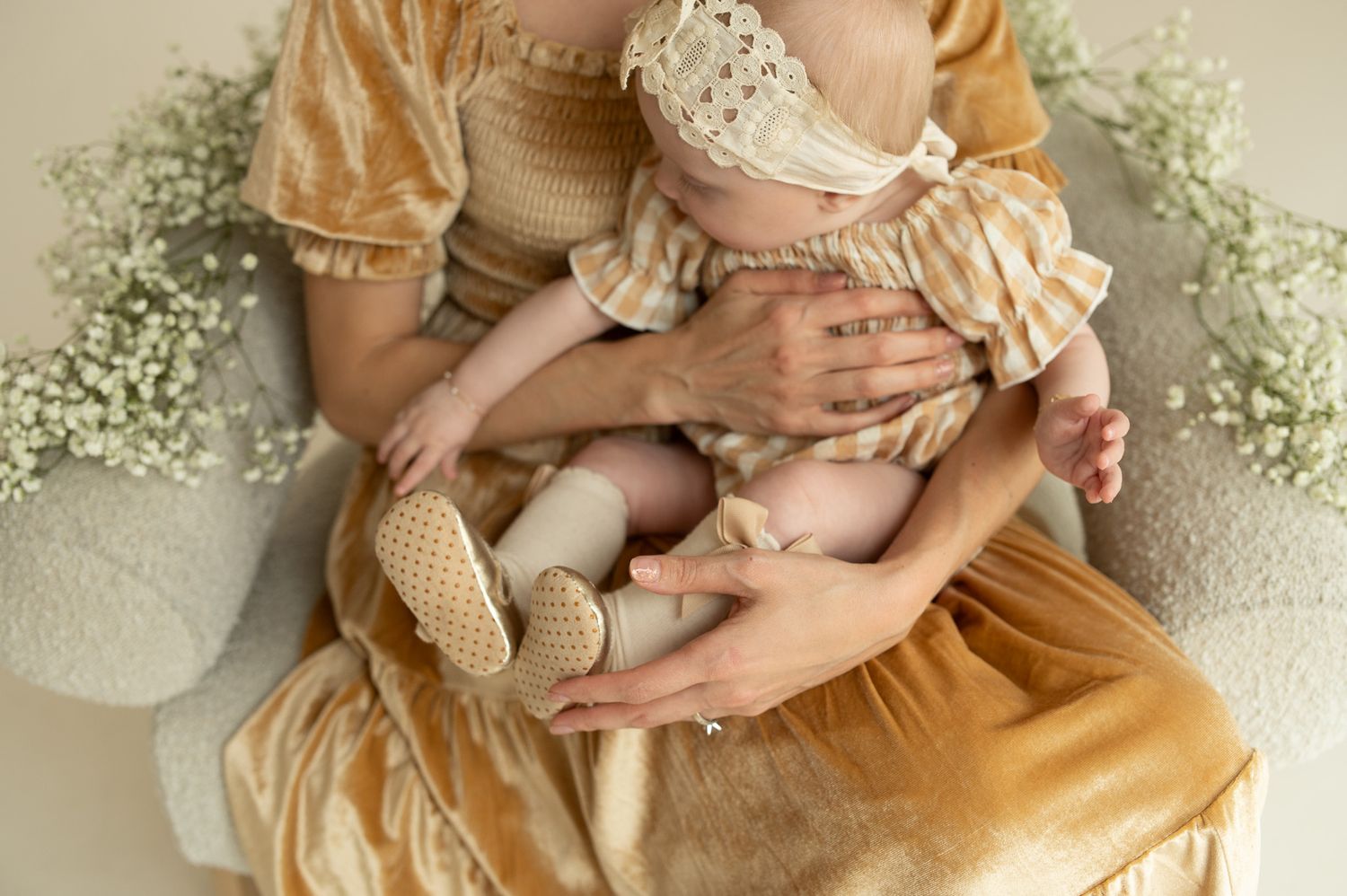 Mother in golden dress cradling newborn baby wearing lace headband, surrounded by white flowers.