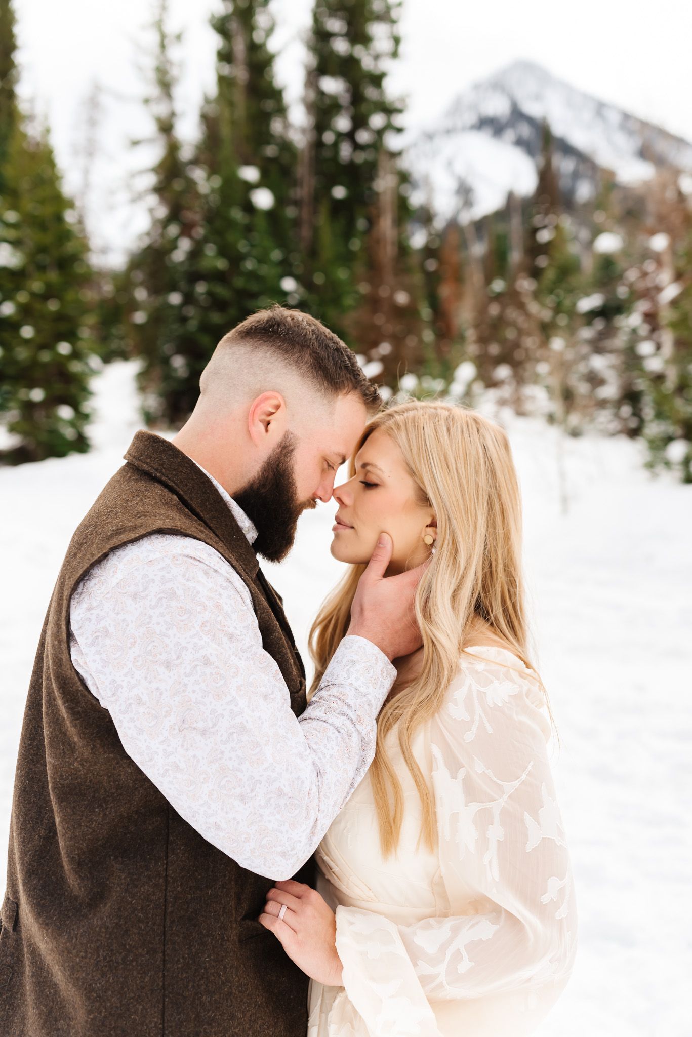 Romantic winter engagement photos in a snowy mountain forest setting with evergreen trees in the background.