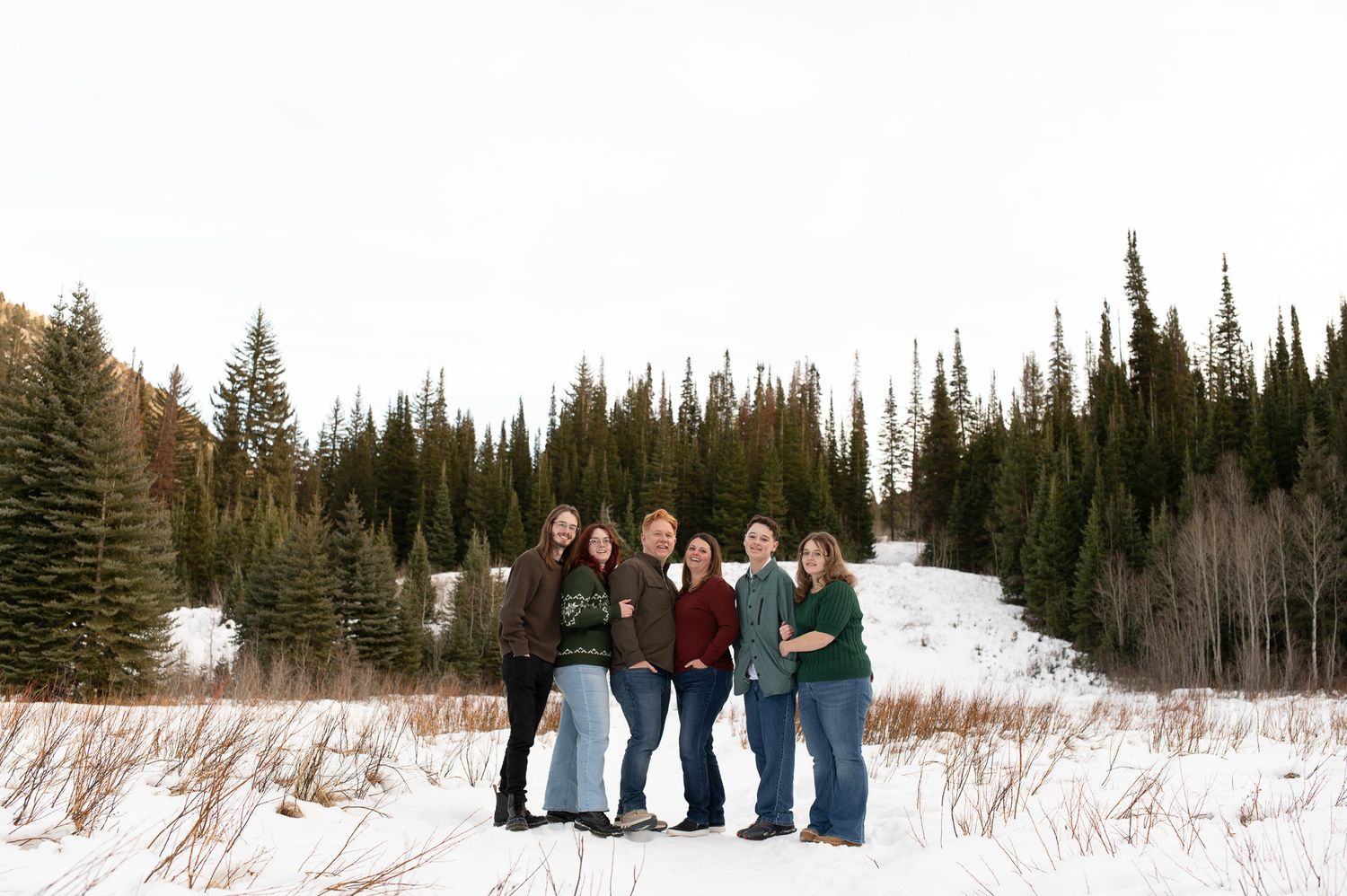 Group of five women standing together in a snowy field surrounded by tall evergreen trees in winter.