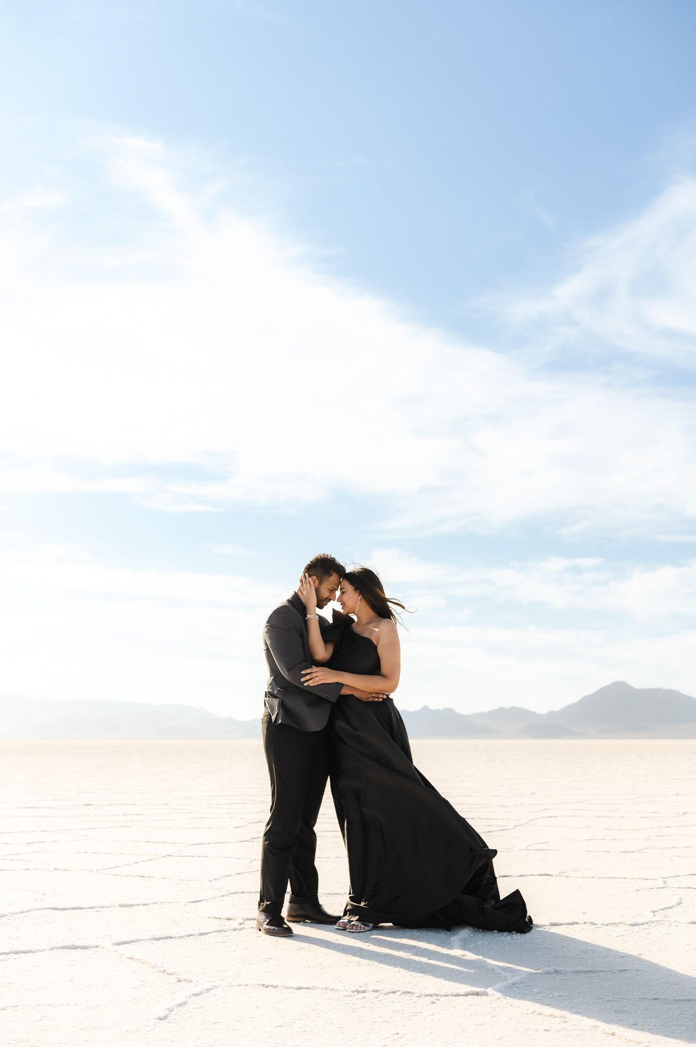 Couple in black clothing embracing on stark white salt flats under bright blue sky.