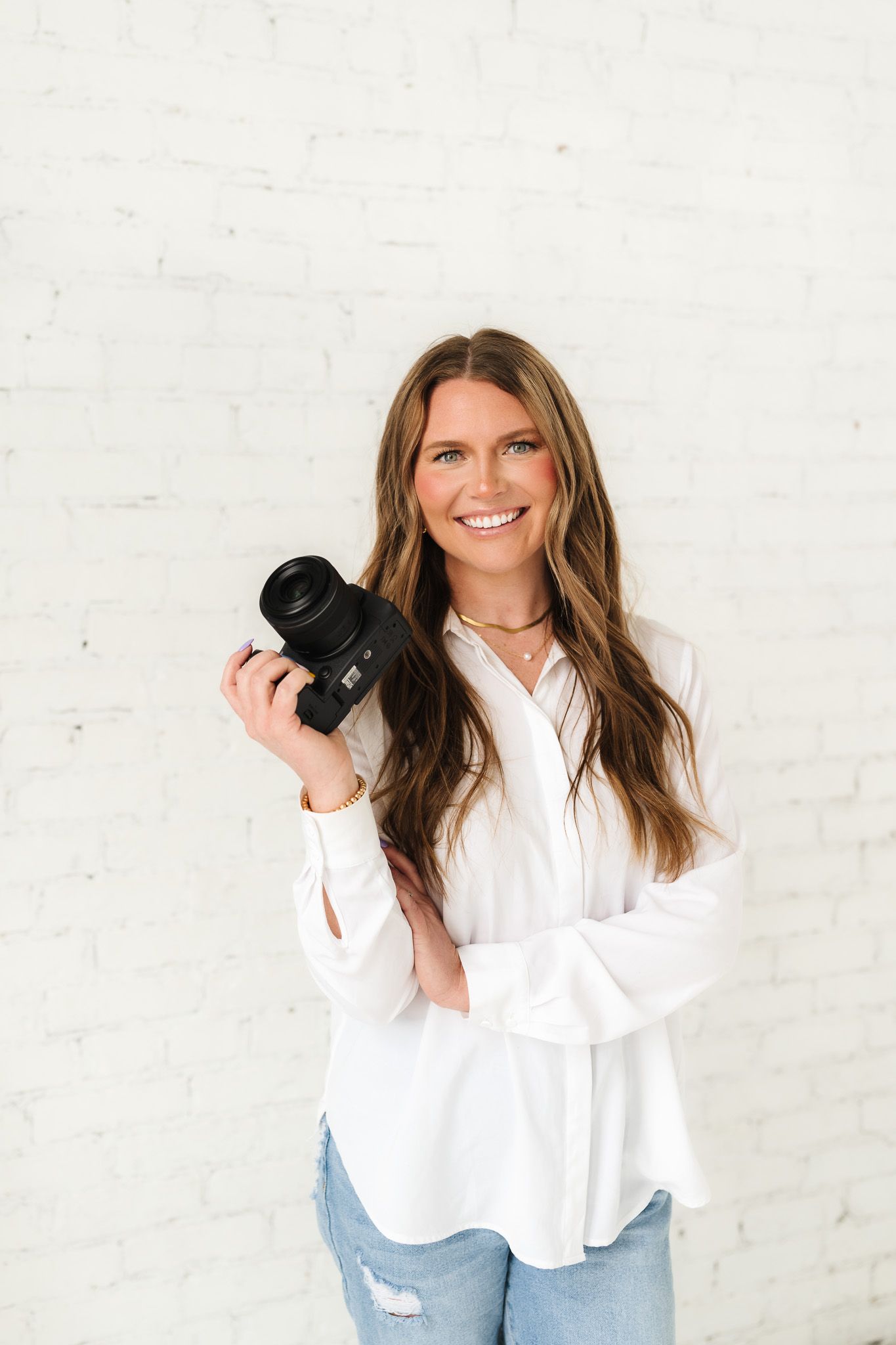Professional photographer in white button-down shirt and jeans holding camera in studio setting.