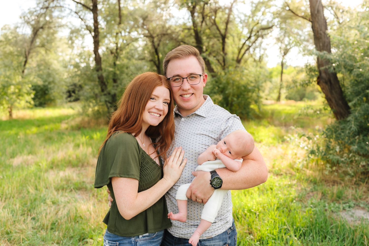 Couple poses with infant during outdoor photography session in green meadow.