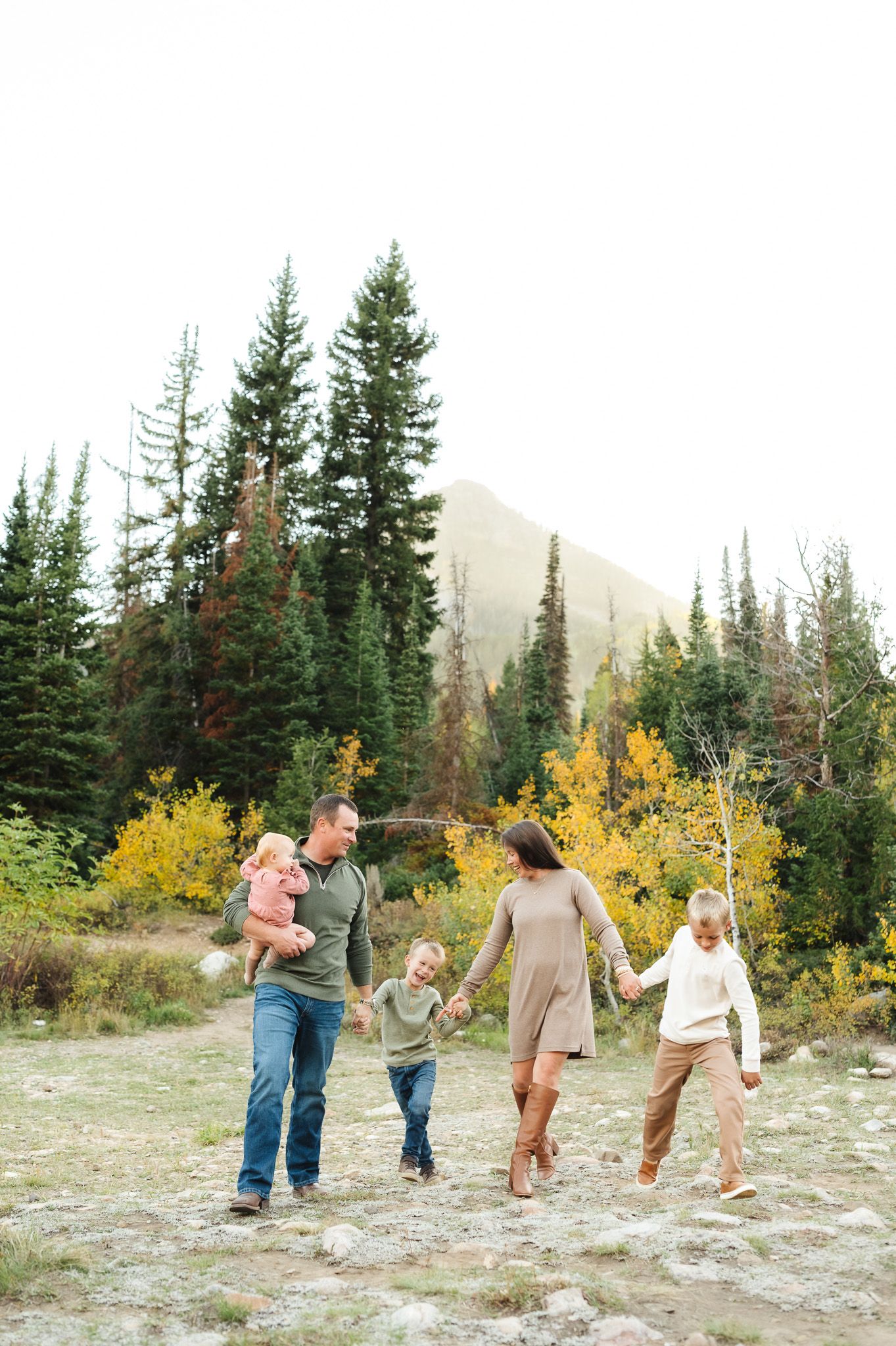 Family holding hands and walking together through a mountain landscape with fall foliage and evergreen trees.